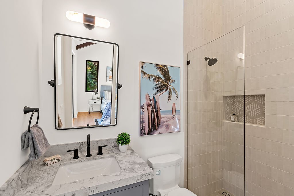 Modern bathroom with a marble vanity, black fixtures, a large mirror, a towel ring holding a towel, a decorative plant, a framed picture of a beach scene, a toilet, and a walk-in shower with beige tiles and a built-in niche.