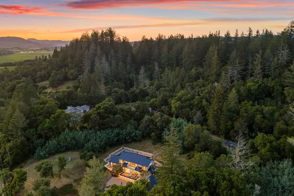 An aerial view of a house in a forested area at sunset, with mountains in the distance and a colorful sky overhead.