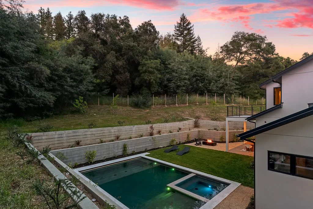 A backyard with a swimming pool and a hot tub, surrounded by a lawn and a wooden privacy fence, with a house to the right, and trees and a colorful sunset sky in the background.