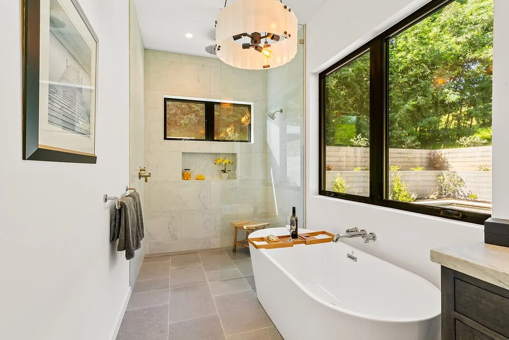 Modern bathroom with a large window showing a garden, a white bathtub with a wooden tray holding a wine bottle, a beige shower area with a small bench, and a decorative light fixture on the ceiling.