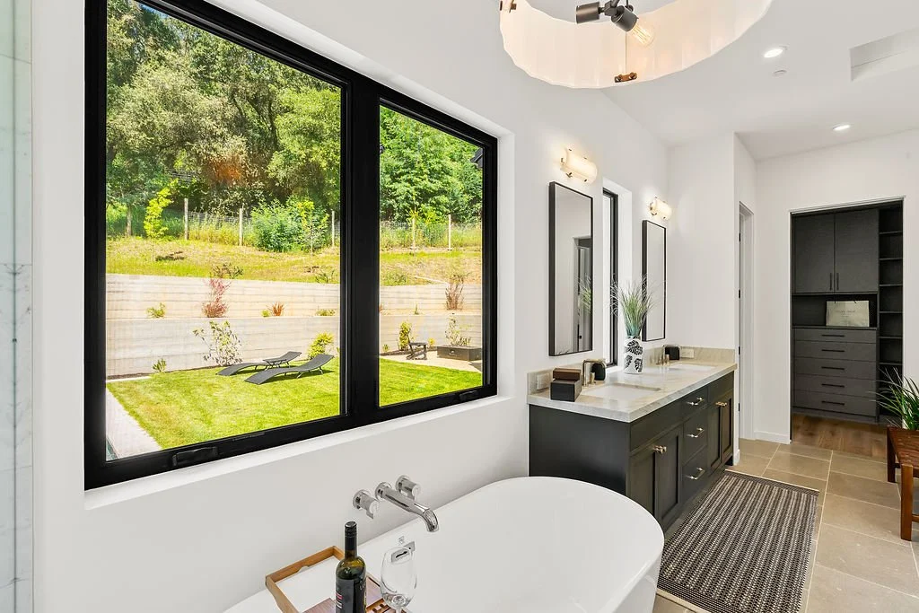 Modern bathroom with a large window overlooking a backyard with grass and lounge chairs, a bathtub, a dual-sink vanity with mirrors, and built-in cabinetry.