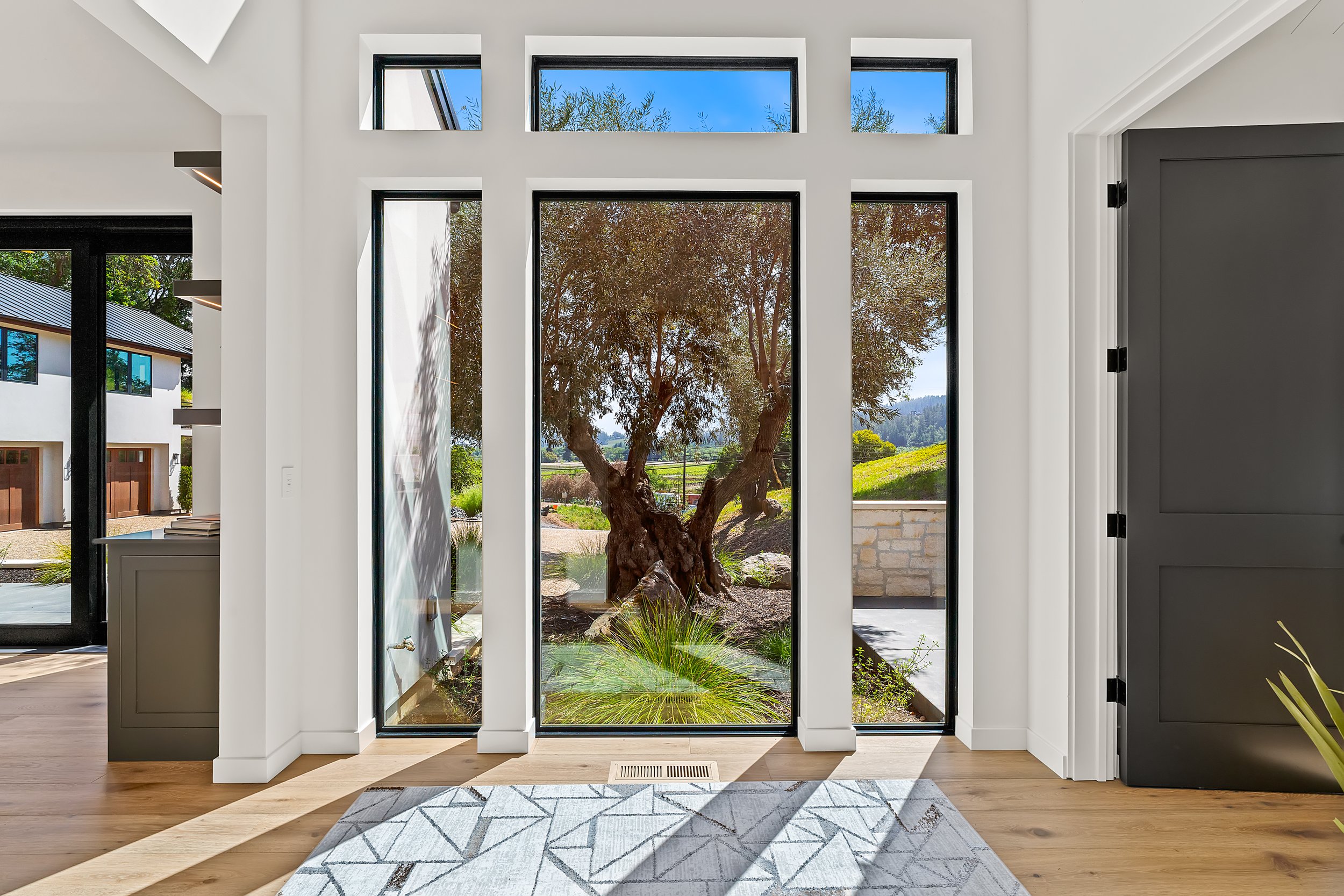 Interior view of a modern home with large floor-to-ceiling glass windows and a glass door, revealing a garden with a tree outside. The room features a light-colored wooden floor, a patterned rug, and a gray cabinet on the right side.