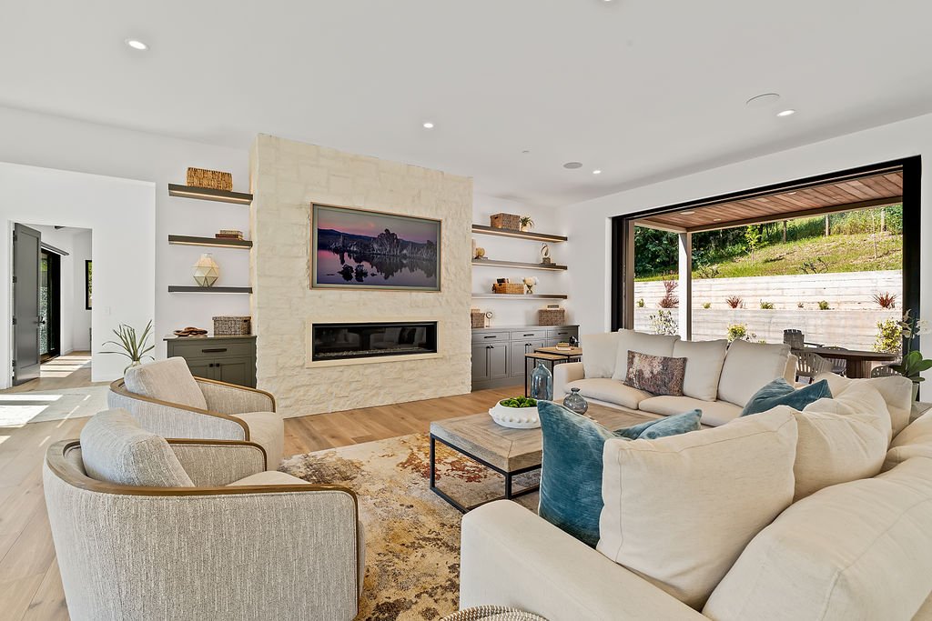 Modern living room with white walls, beige furniture, a stone fireplace with a television, wooden floors, and large windows overlooking a garden.