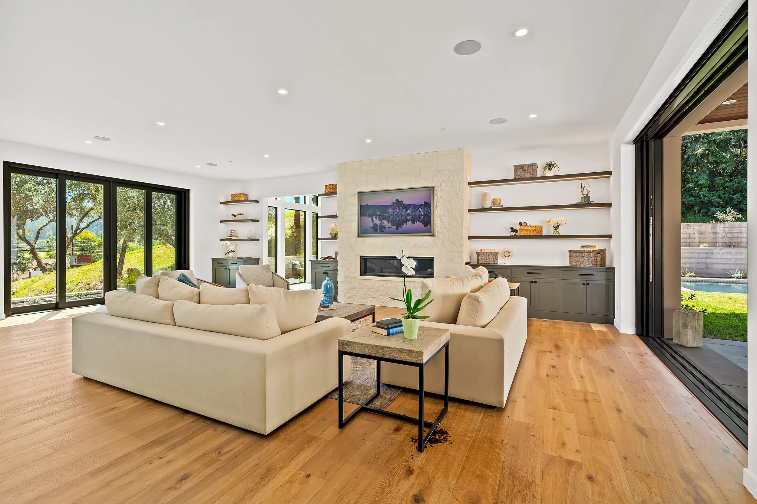 Spacious, modern living room with large sliding glass doors opening to a lush green outdoor view, beige sofas, a wall-mounted TV, and built-in shelves with decorative items.