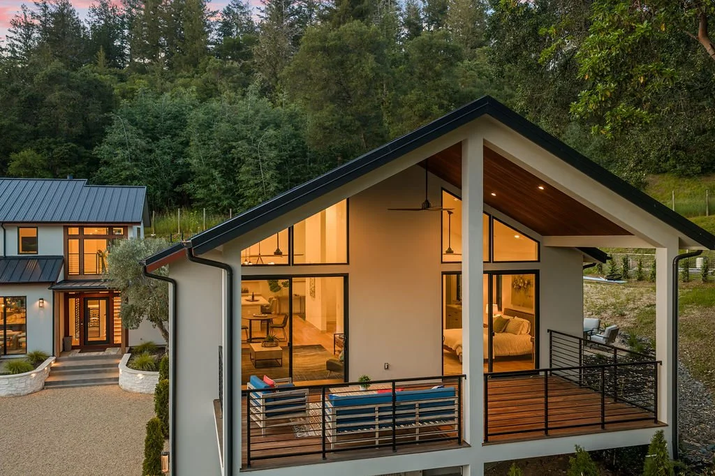 A modern house with large glass windows and a balcony, surrounded by trees, evening lighting inside showcasing living and dining areas.