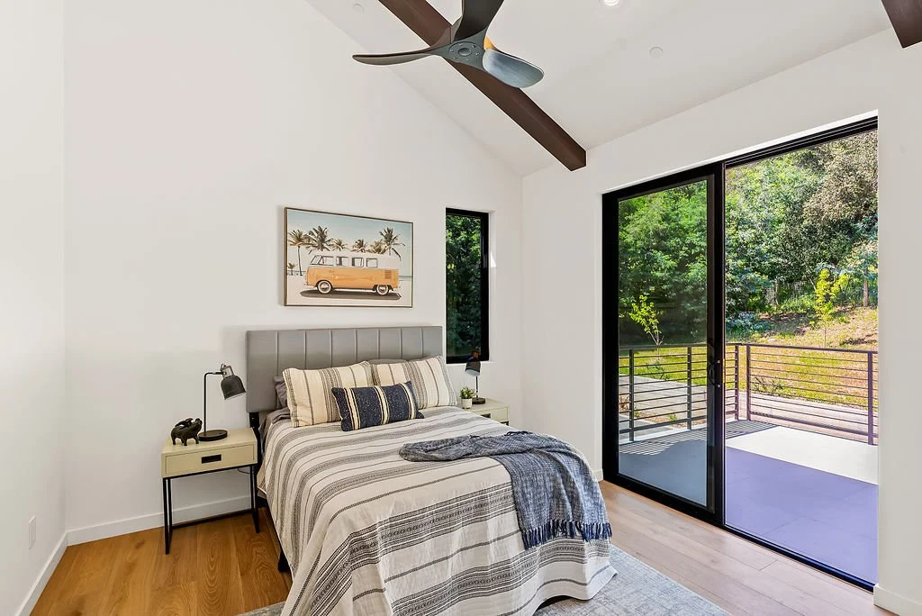 A modern bedroom with a bed, nightstands, and large sliding glass doors opening to a balcony, overlooking a lush green outdoor area.
