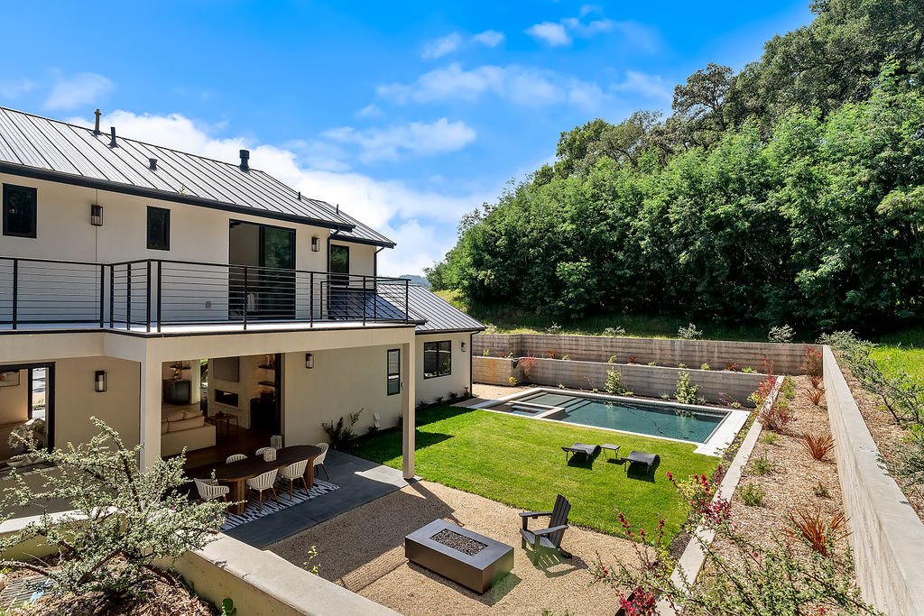 Modern two-story house with a backyard pool, outdoor dining area, and view of green hillside.