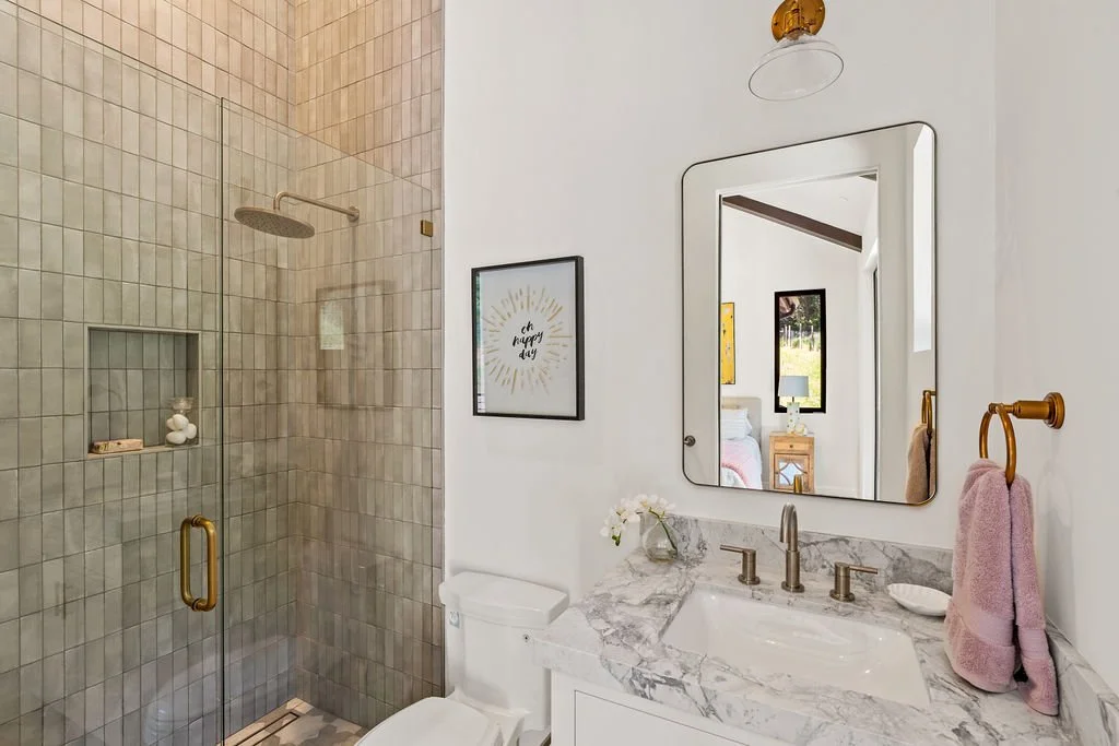 Bathroom with a glass shower enclosure, marble countertop sink, large wall mirror, framed decorative art, pink towel, small vase with flowers, and a view of a bedroom with a table lamp and window.