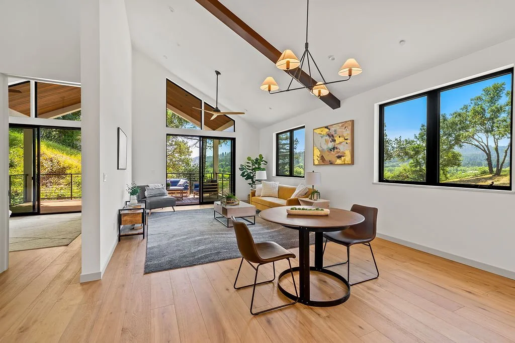 Modern living room with large windows overlooking greenery, a gray rug, a yellow sofa with white and beige pillows, a small gray armchair, a round dining table with three brown chairs, a ceiling light fixture, and a sliding glass door leading to a pa