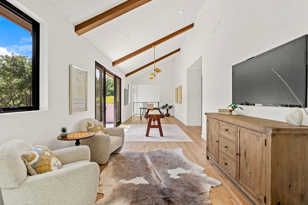 Living room with white walls, wooden ceiling beams, a large flat-screen TV on a wooden cabinet, two beige armchairs with decorative pillows, a cowhide rug, windows and a sliding glass door bringing in natural light, and a view of trees outside.