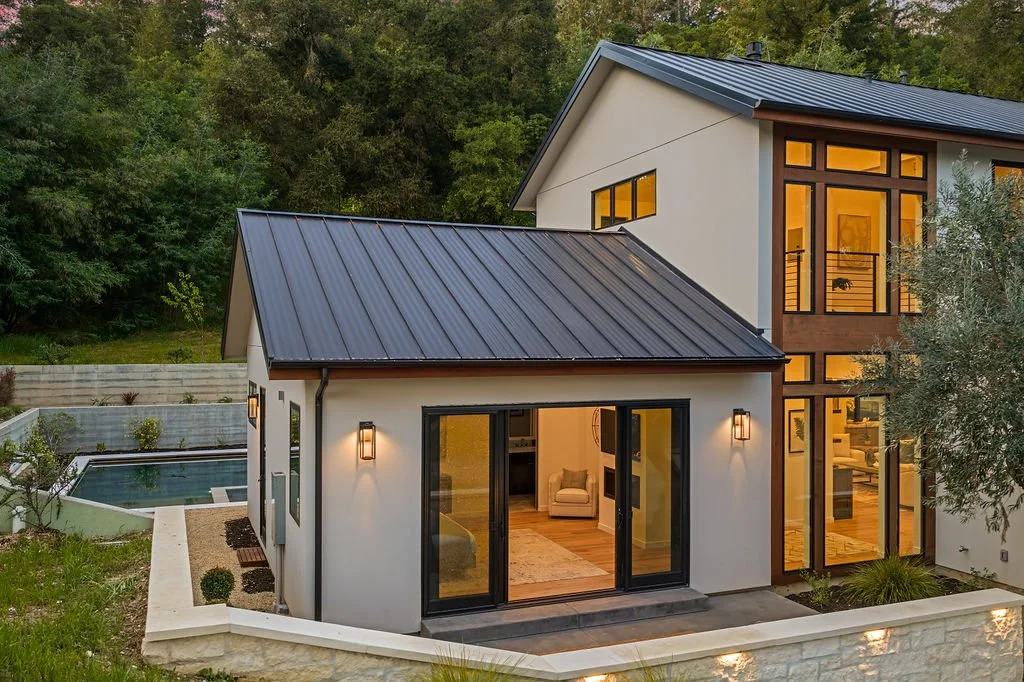Modern two-story house with large glass windows and sliding doors, surrounded by a yard with a pool and greenery, during evening hours.