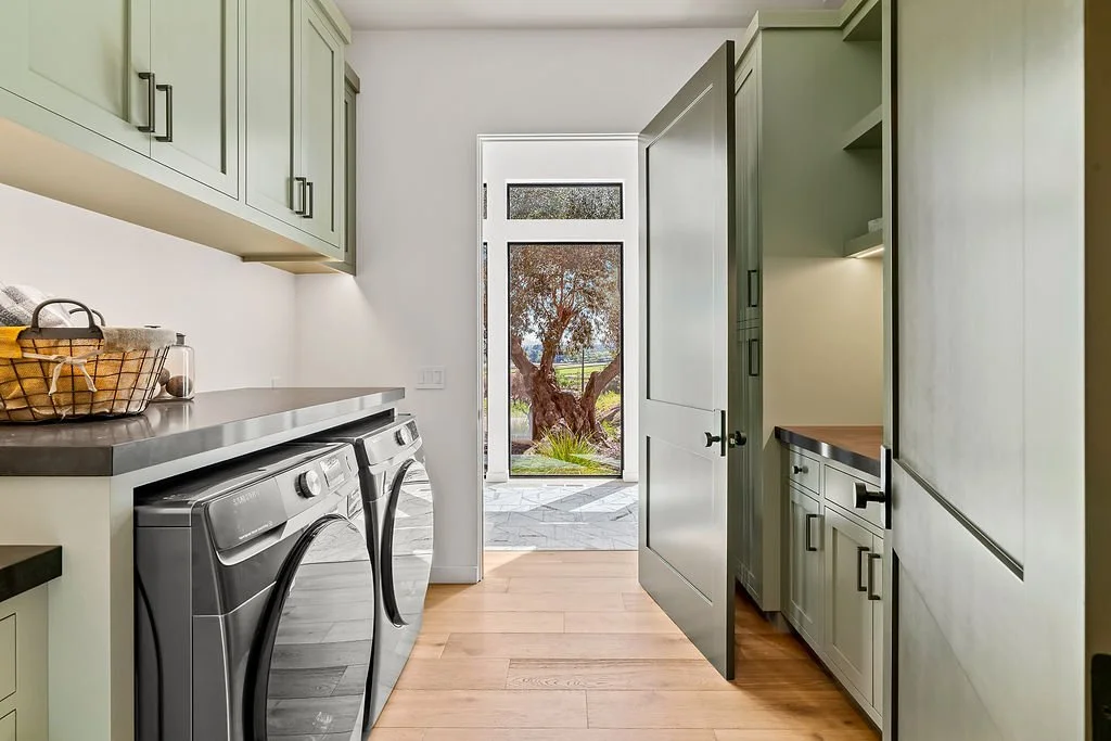 Laundry room with light green cabinets, a washer and dryer, and an open door leading outside to a patio and tree.
