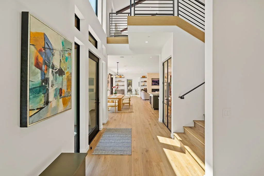 Bright modern hallway with wooden flooring, abstract wall art, staircase, and view into living and dining areas.