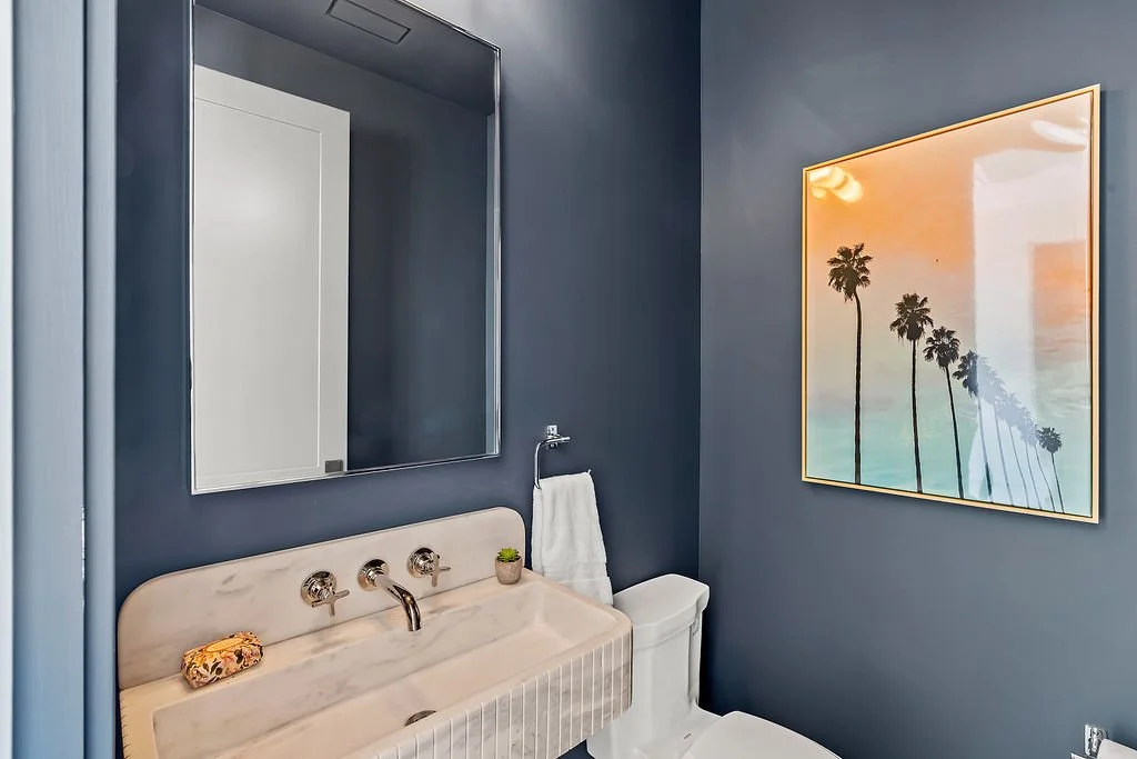 A bathroom with dark blue walls, a large mirror, a white marble sink with three taps, a small potted plant, a white towel on a ring, and a framed picture of palm trees at sunset.