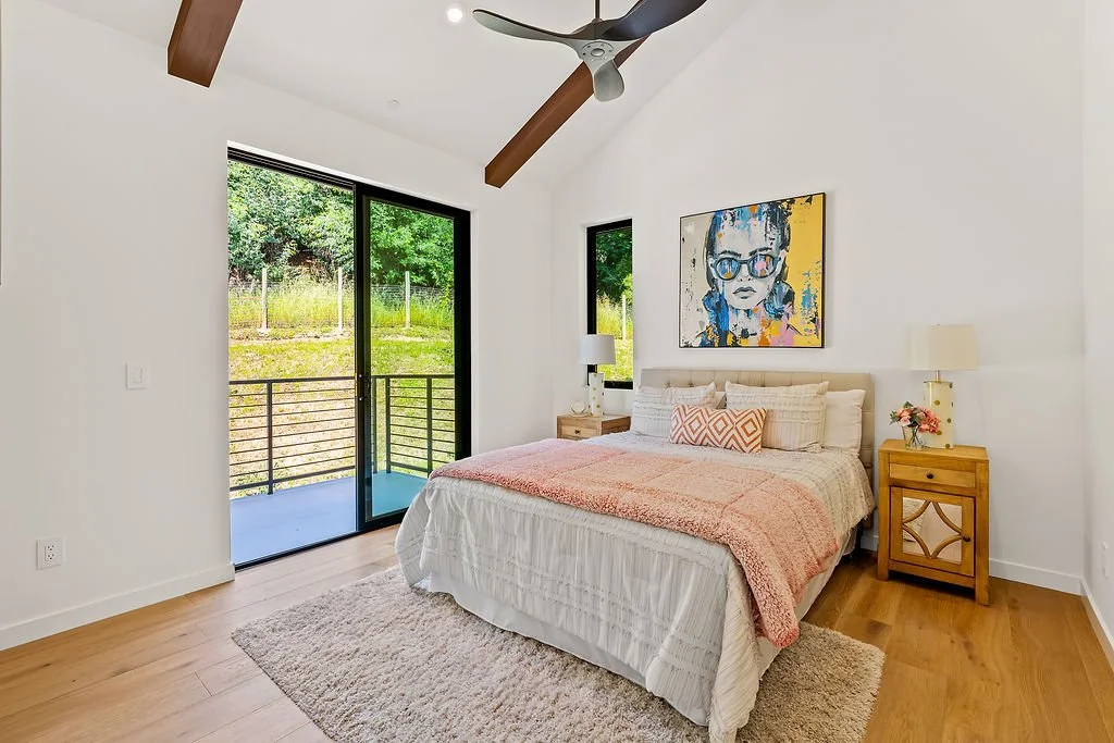 A bedroom with a large bed covered in white and pink bedding, two wooden nightstands with lamps, a colorful artwork of a girl on the wall, a sliding glass door opening to a small balcony overlooking greenery, a window next to it, wood flooring, ceili