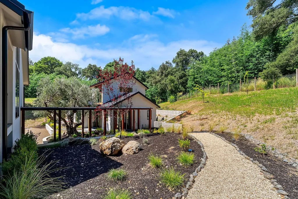 A backyard garden with a gravel pathway, some rocks, plants, and a small tree next to a house with a deck. In the background, there are more trees and a blue sky with clouds.