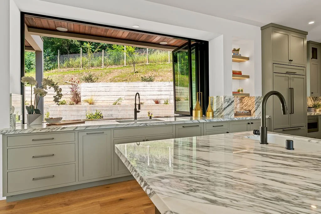 Modern kitchen with marble countertops, gray cabinets, a large open window overlooking a landscaped backyard, and decorative vases.