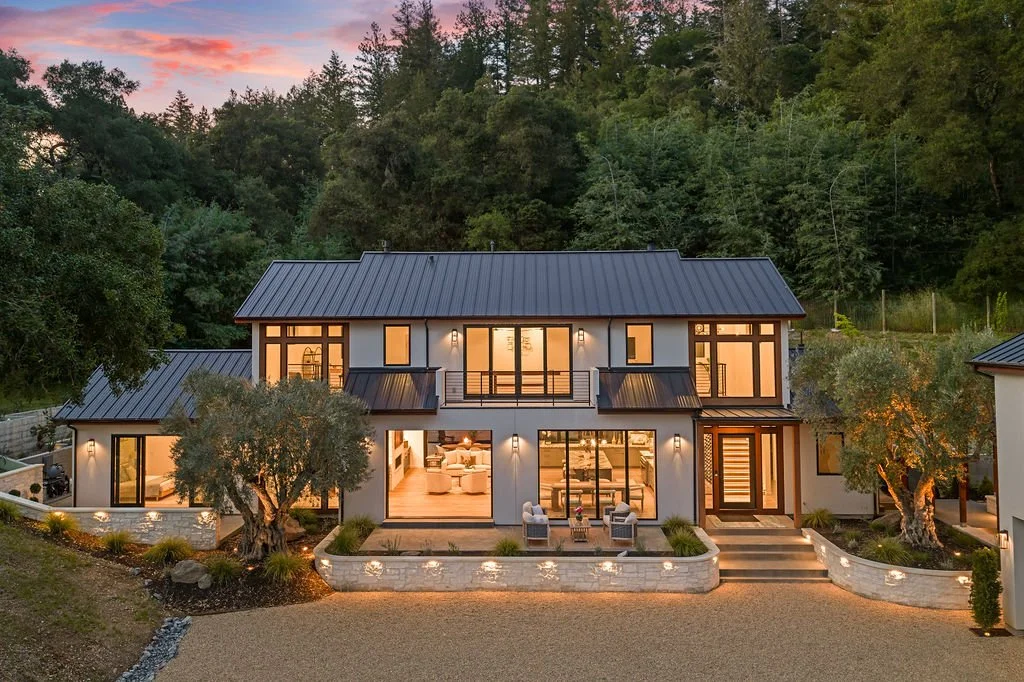 Modern two-story house with large windows, located in a wooded area at dusk, with outdoor lighting and a small seating area in front.