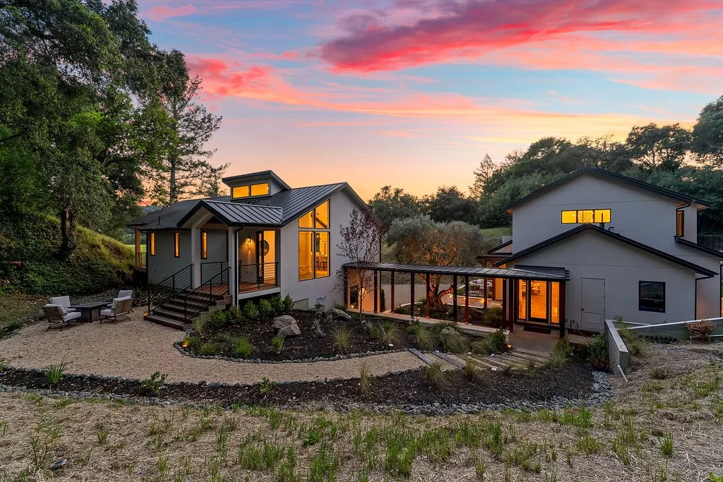 Modern two-story house with large windows at sunset, surrounded by trees and a landscaped yard with gravel pathways and outdoor seating.