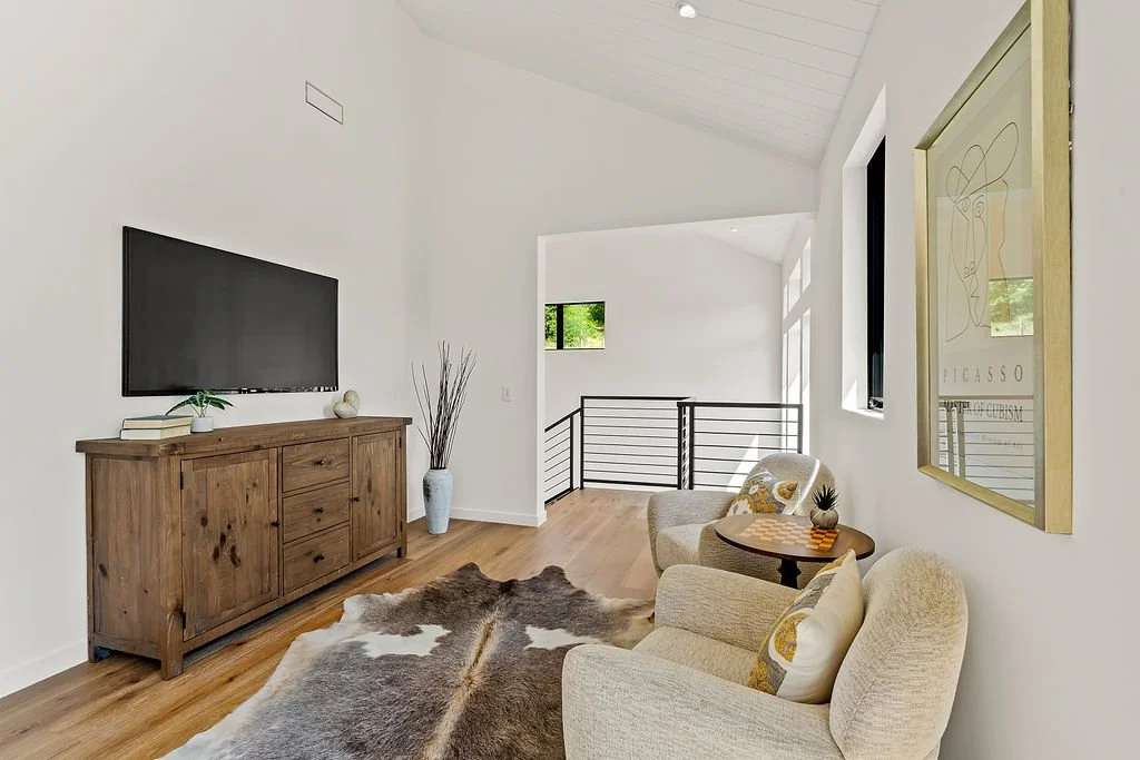 Modern living room with white walls, wooden furniture, seating area with two beige chairs and a small wooden side table, a cowhide rug, framed artwork, and large windows with natural light.