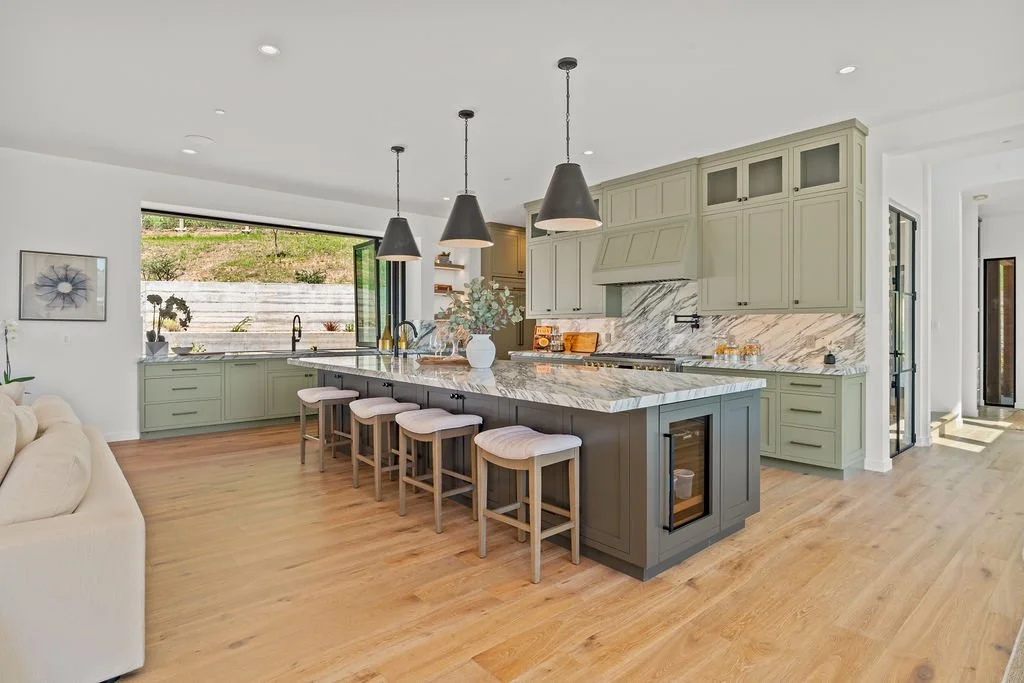 Modern kitchen with light green cabinets, marble countertops, and pendant lighting over a kitchen island with four chairs. Open-plan space with hardwood flooring and a large window.