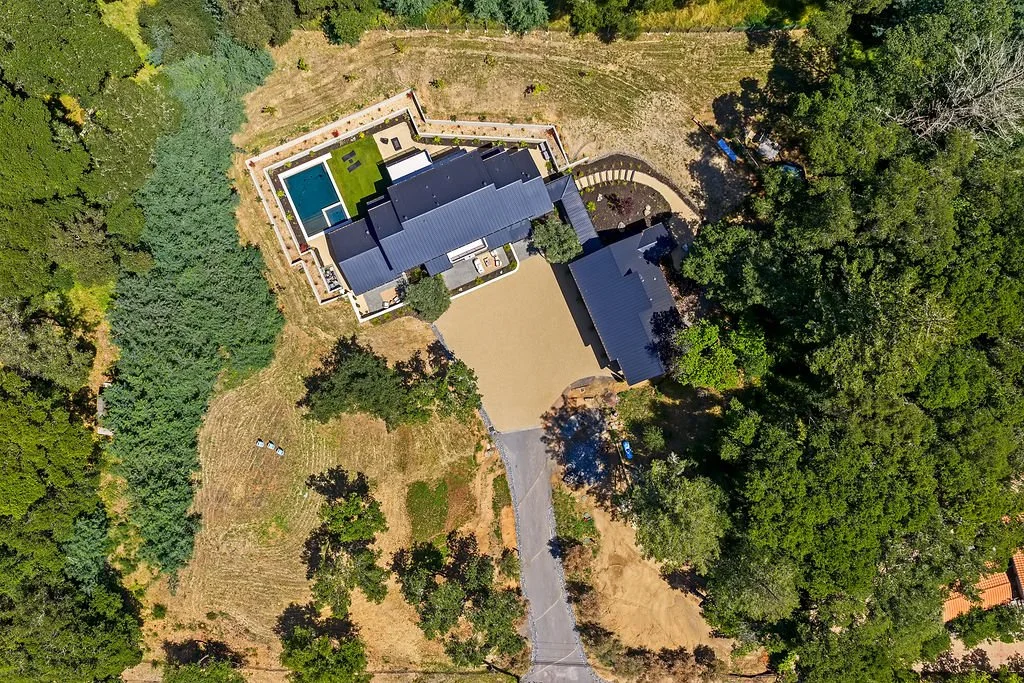 Aerial view of a large house with a blue metal roof, surrounded by trees and a spacious yard with a driveway, swimming pool, and patio area.