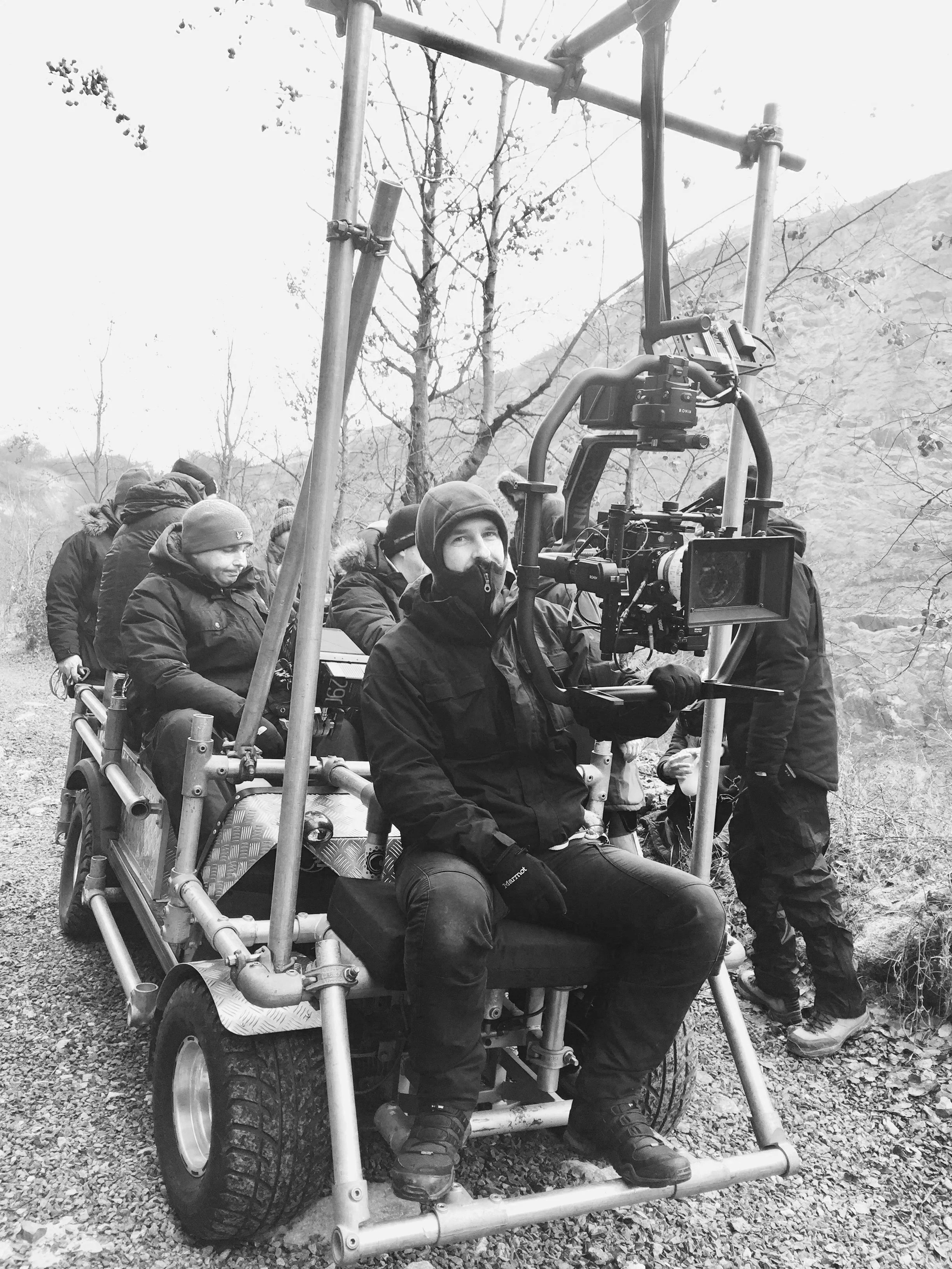 Black and white photo of a film crew operating a camera on a DJI ronin 2 3 axis stabiliser outdoors in a wooded area, with several crew members dressed in winter clothing.