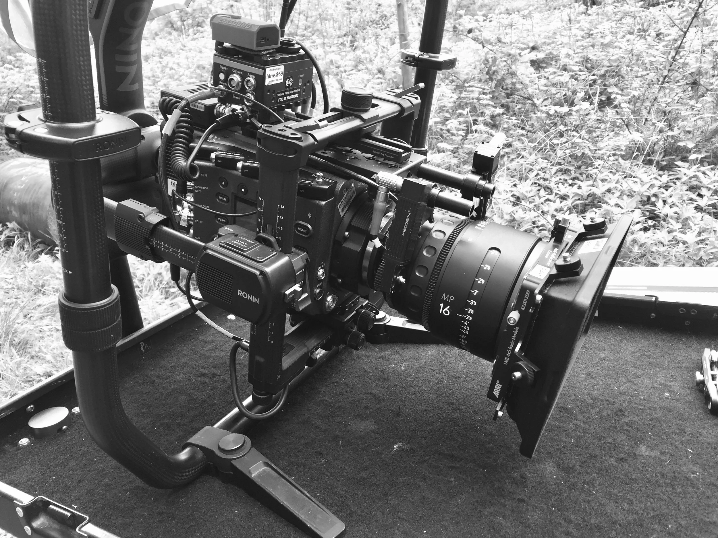 A professional video camera setup situated outdoors with various attached equipment, on a surface with grass and foliage in the background.