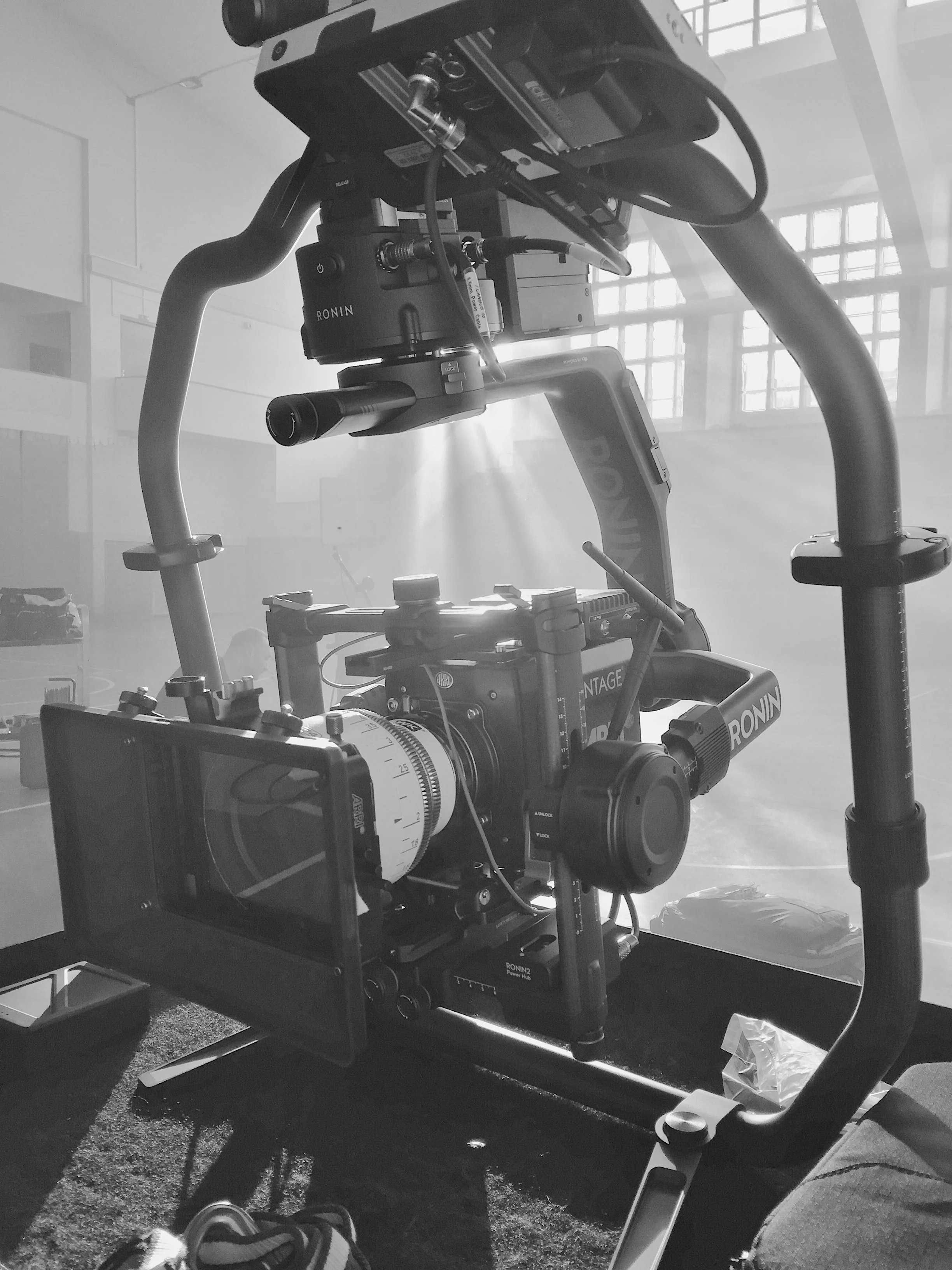A professional video camera mounted on a sturdy tripod in a large indoor space with high ceilings and grid windows, likely used for filming or broadcasting.
