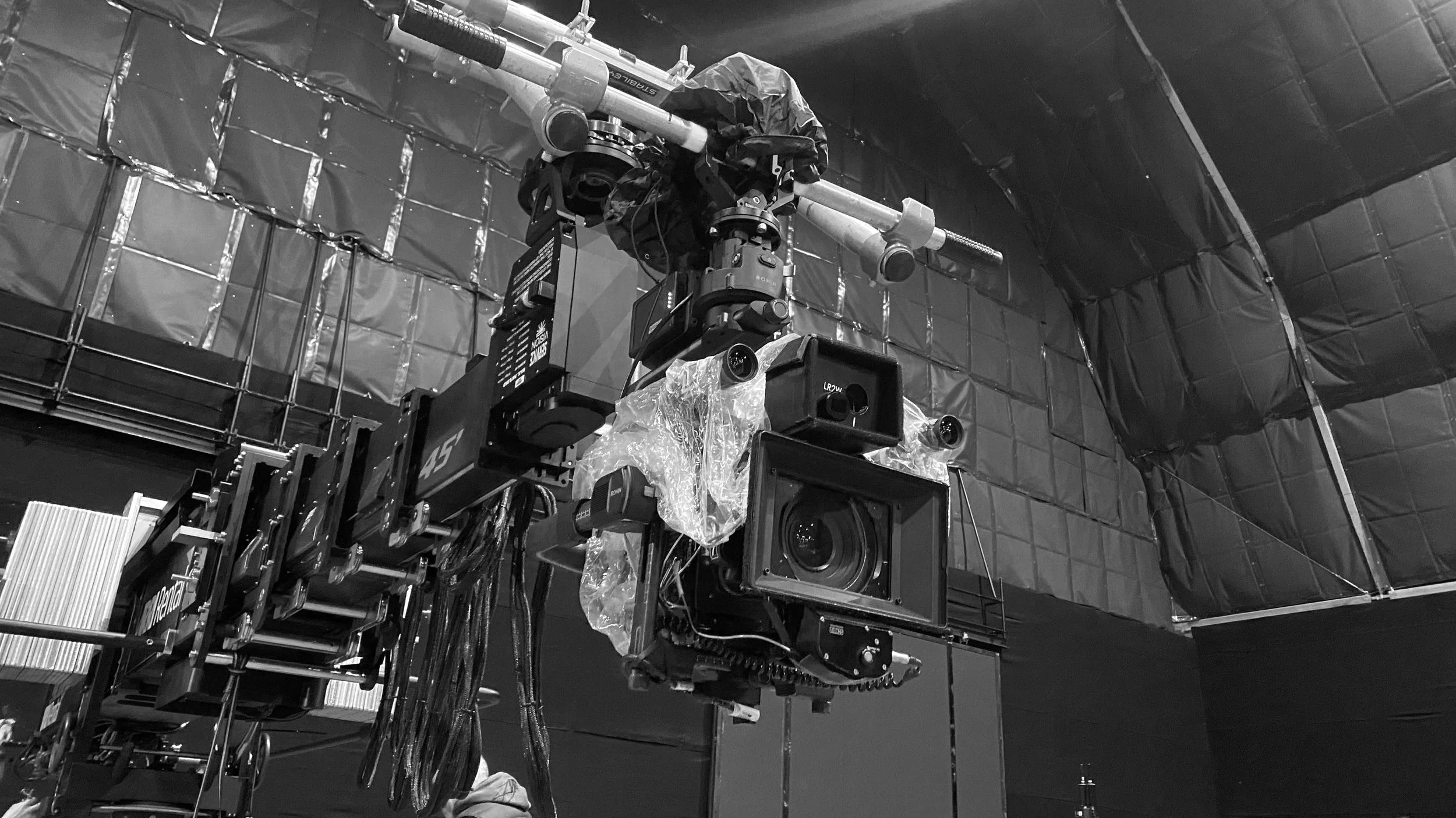 A large industrial camera or sensor mounted on a sturdy metal frame, with a black protective cover around its lens, situated inside a dark room with reflective insulation on the walls.
