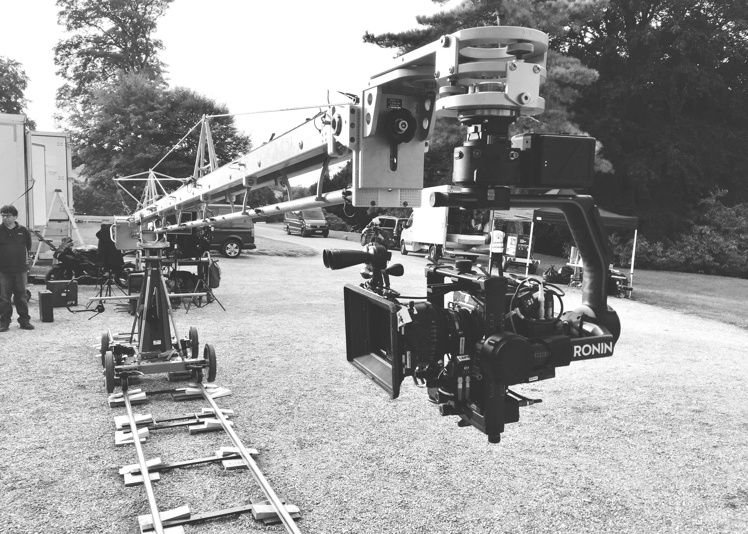 A film production scene with a large camera mounted on a crane, and crew members near parked vehicles and equipment outdoors.