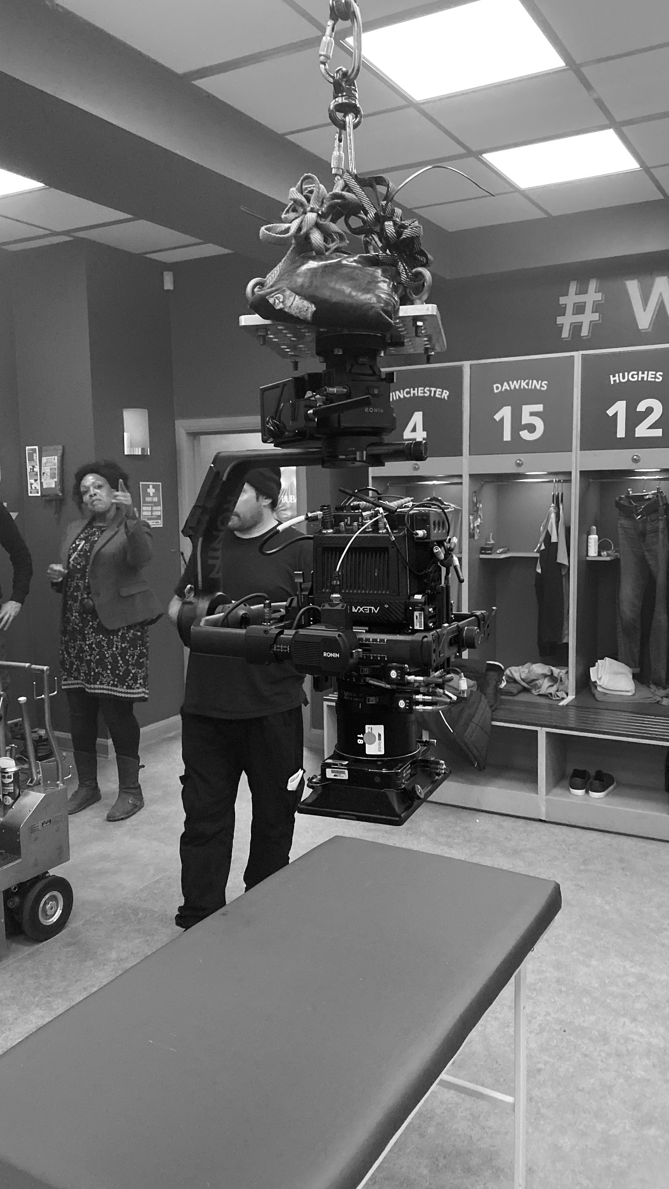 Black and white photo of a film or television set with a camera crane, crew members, and a locker room in the background.