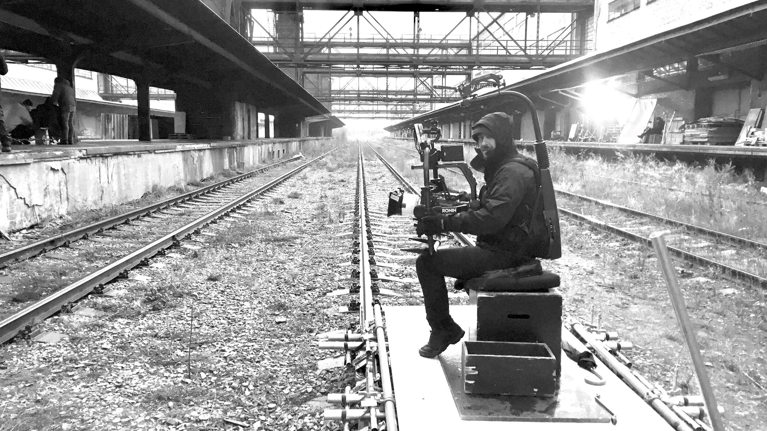 A person operating a camera on a track in a train station or industrial setting, with railway tracks on either side and structures overhead, in black and white.