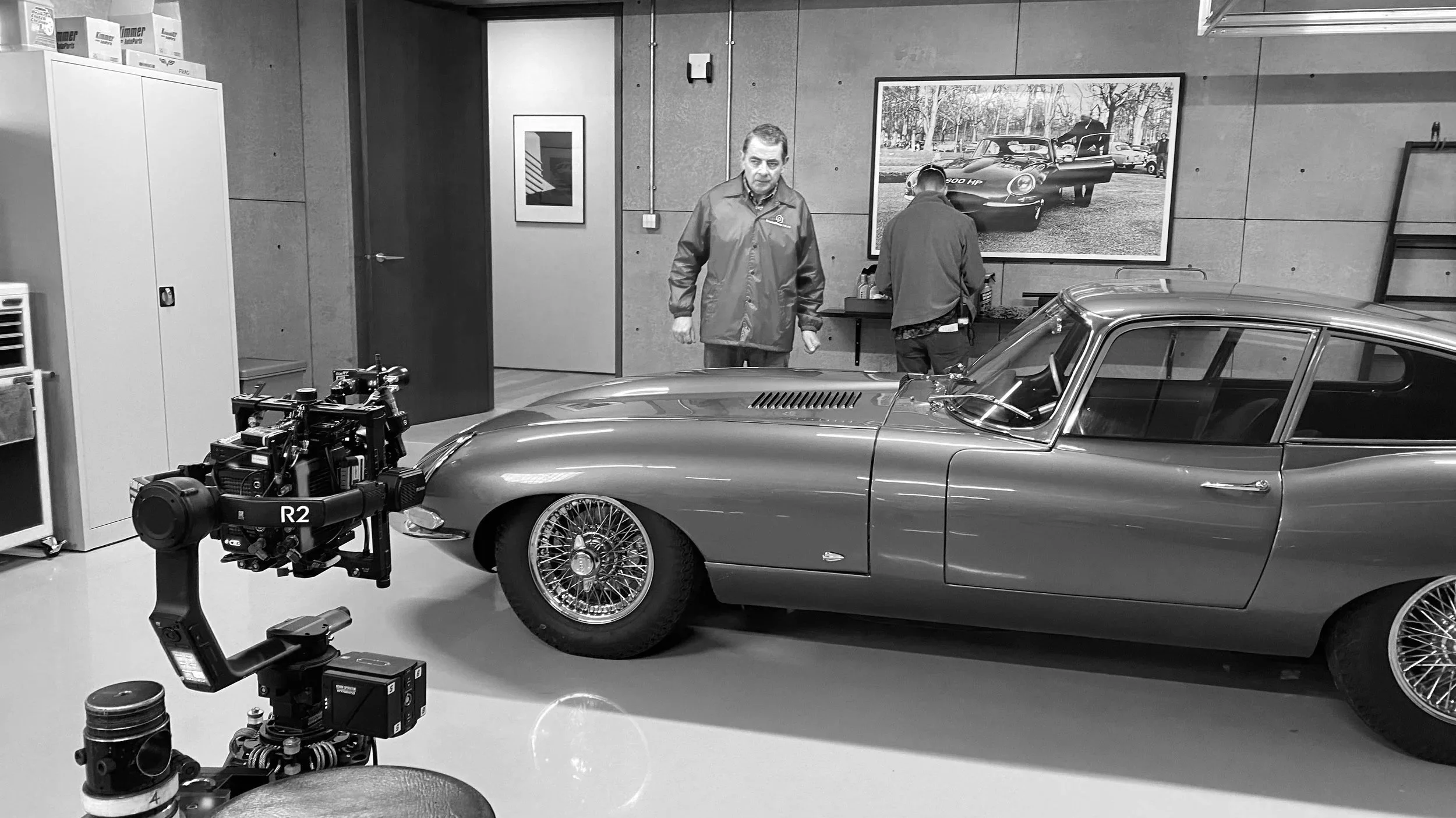Black-and-white photo of a vintage sports car in a garage with two men nearby, one inspecting and the other with his back turned; a professional camera rig in the foreground.