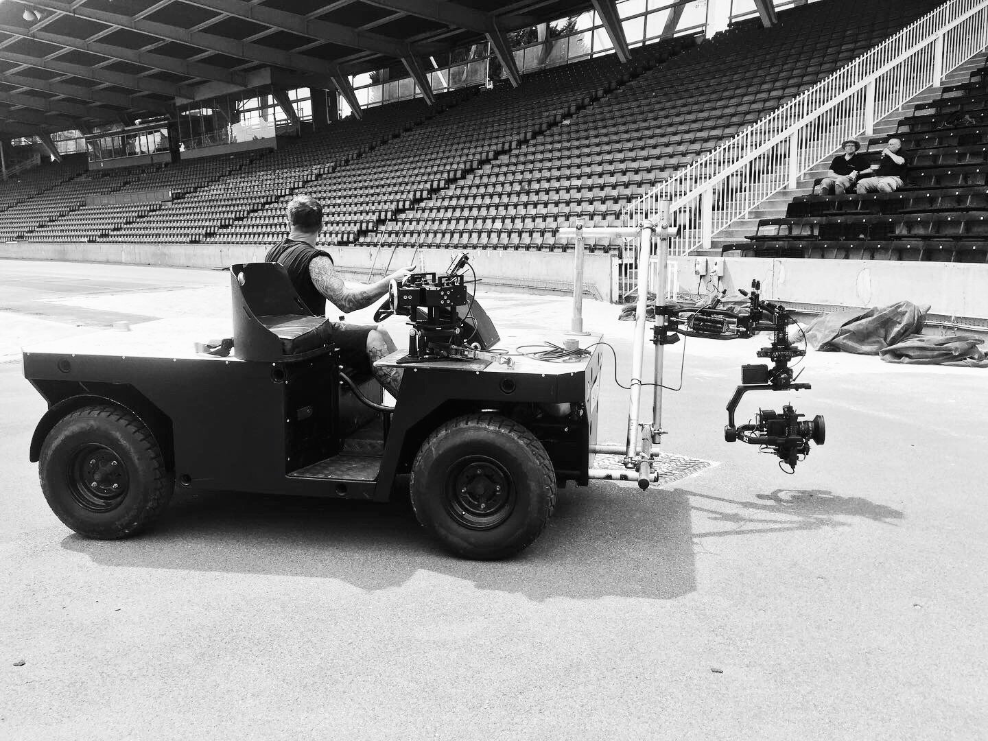 Black-and-white photo of a filming crew on a deserted sports stadium field, with a camera mounted on a small off-road vehicle and two people seated in the stands.