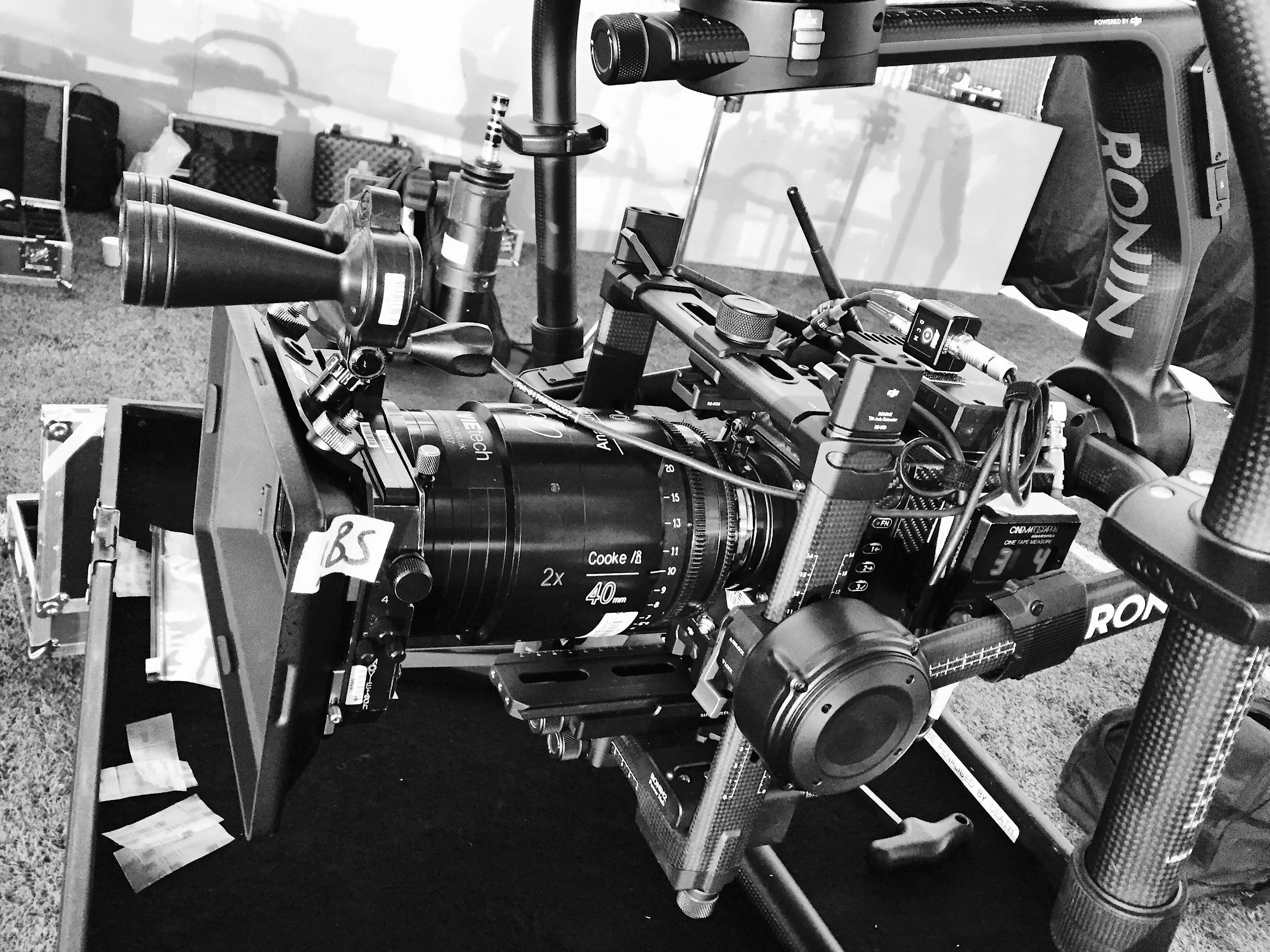 Black and white photograph of a professional film camera on a DJI ronin 2, surrounded by equipment cases and cables, in an outdoor setting.