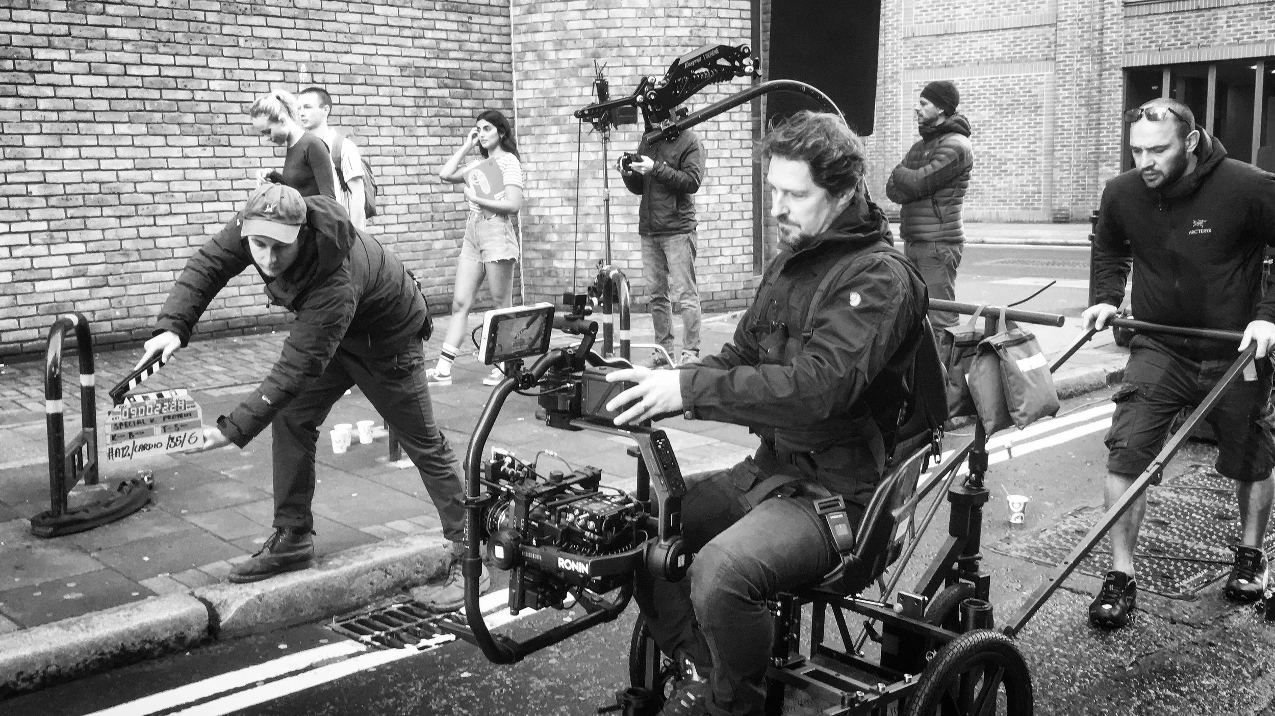 Film crew set up on city street with camera equipment, crew members preparing for shoot, brick buildings in the background, black and white photograph.