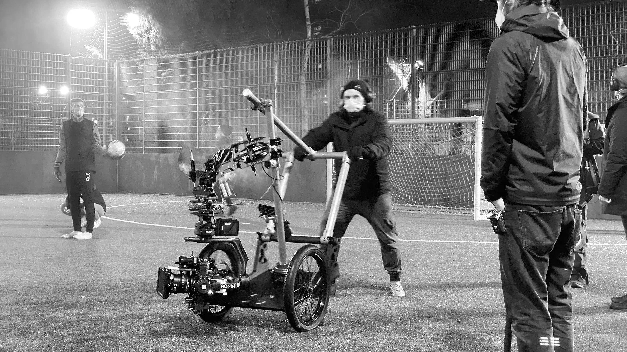A group of people on a nighttime outdoor sports field, with one person operating a camera mounted on a small electric vehicle, and others standing or walking nearby, all wearing masks and warm clothing.