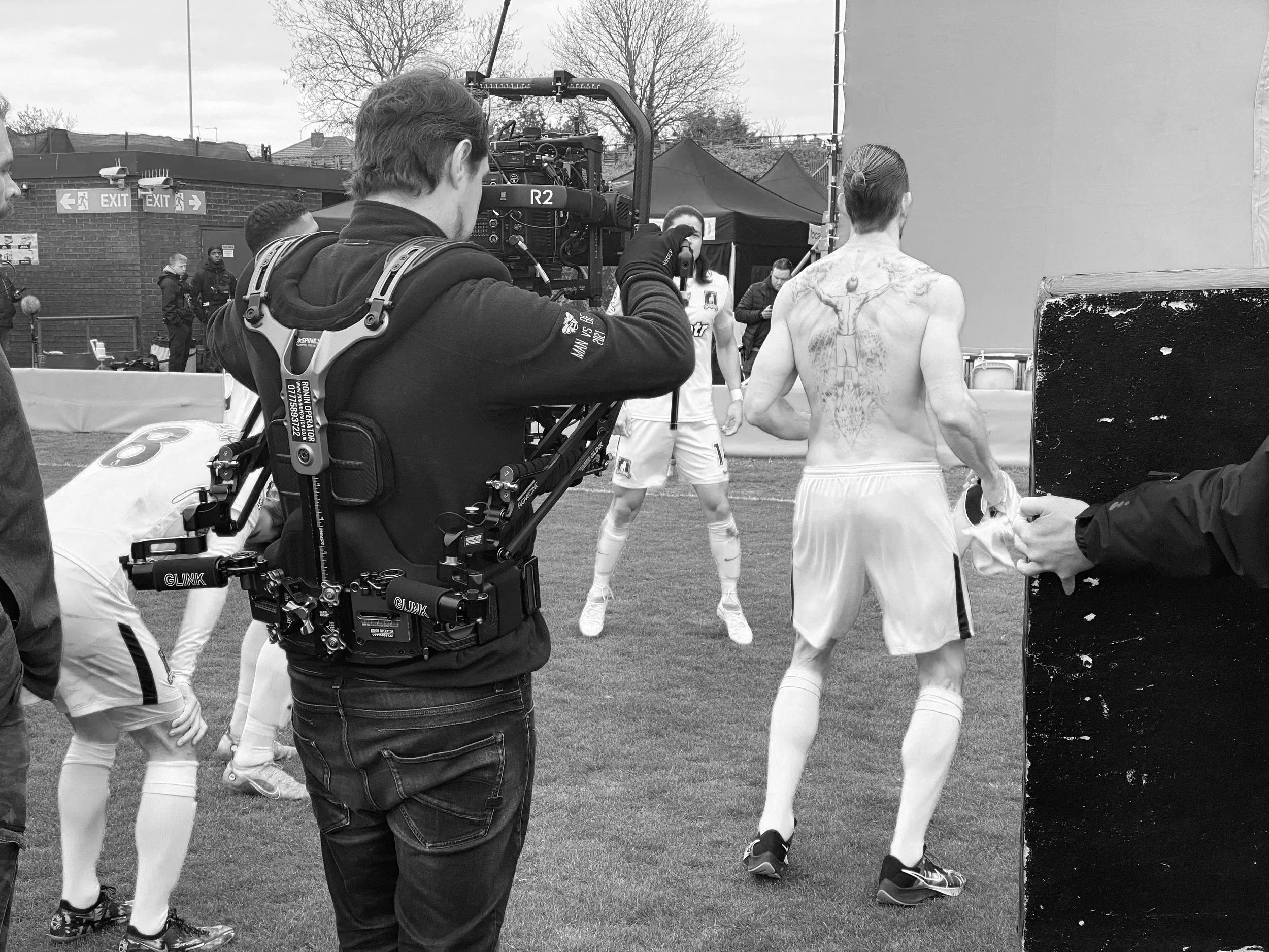 Black and white photo of a film crew filming a sports event with a camera operator, wearing a backpack, filming a shirtless athlete with a large tattoo on his back and wearing sports gear, on an outdoor field with trees in the background.