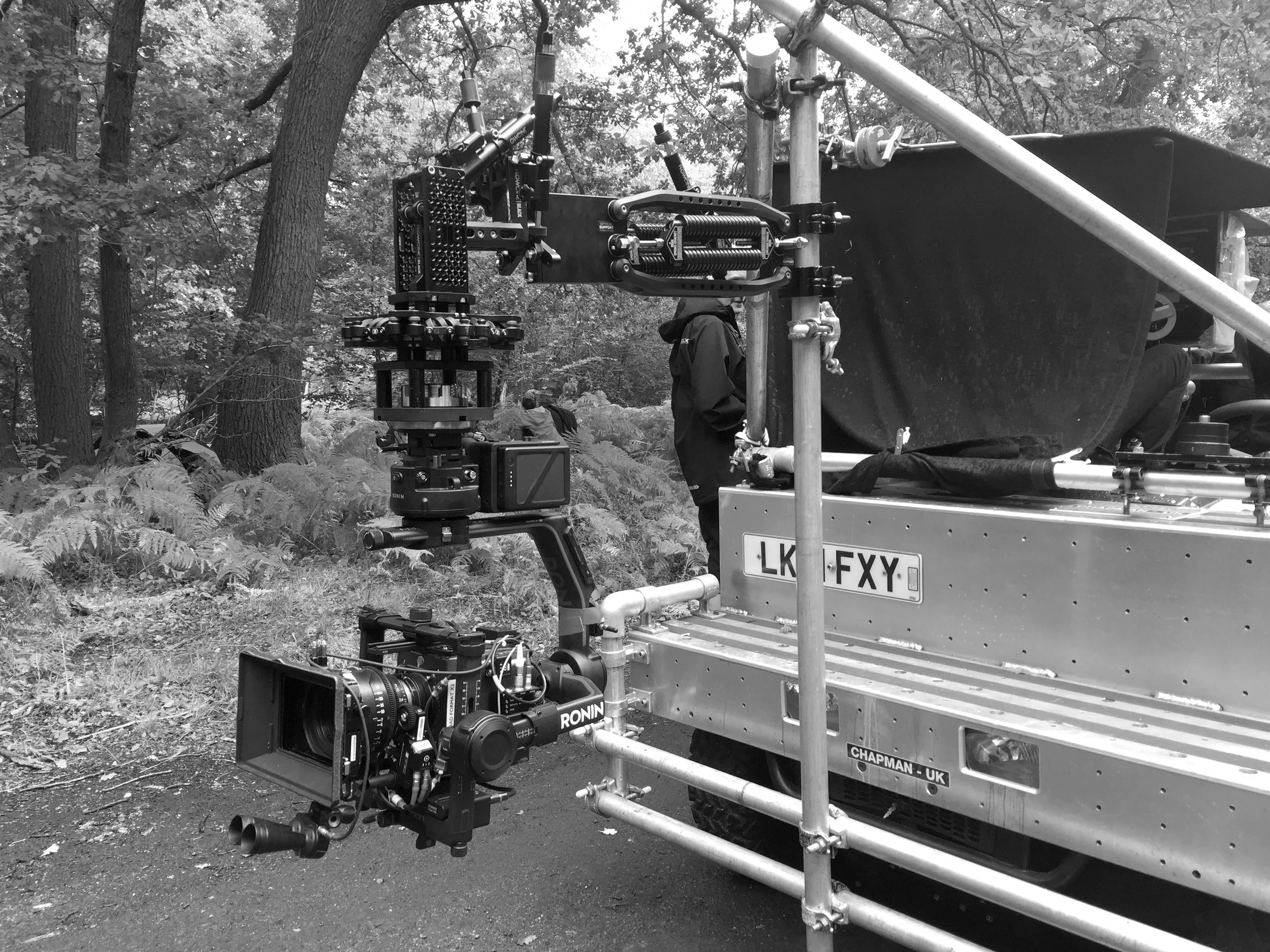 A professional movie or television camera mounted on a Flowcine black arm, with a film crew set up outdoors in a wooded area. The scene includes metal scaffolding and filming equipment, with trees and foliage in the background.