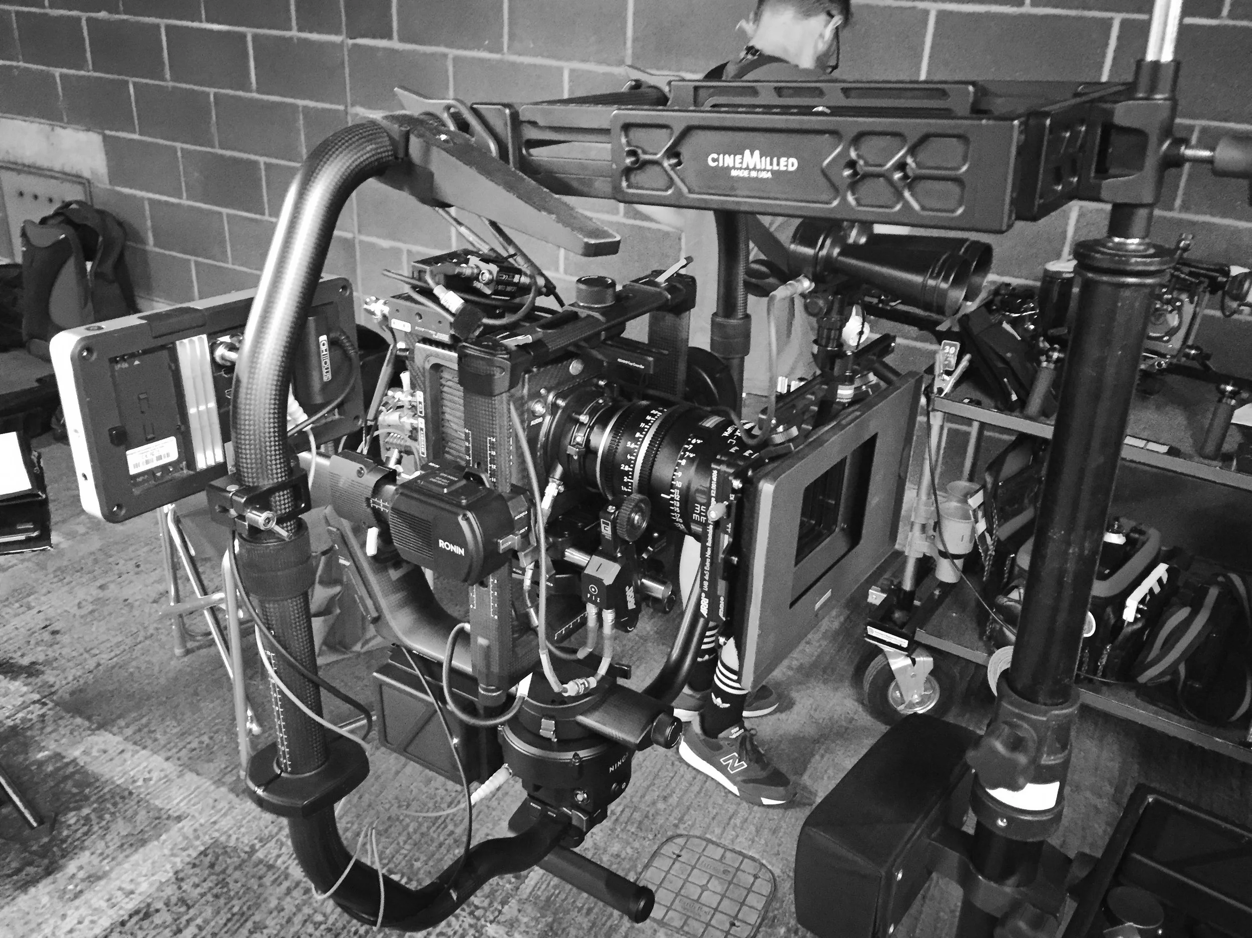 A professional film camera setup with various accessories on a cart, including a monitor, cables, and other filmmaking equipment, against a brick wall.