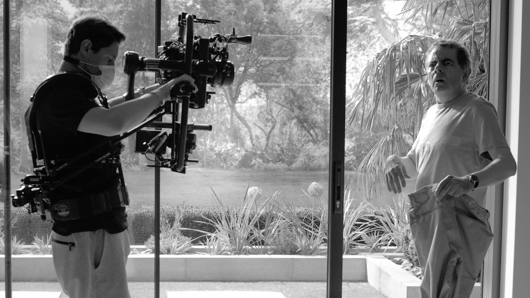 Behind the scenes of a film shoot showing a cameraman operating a 3 axis stabiliser rig and a man holding his trousers inside a house near large windows and plants.