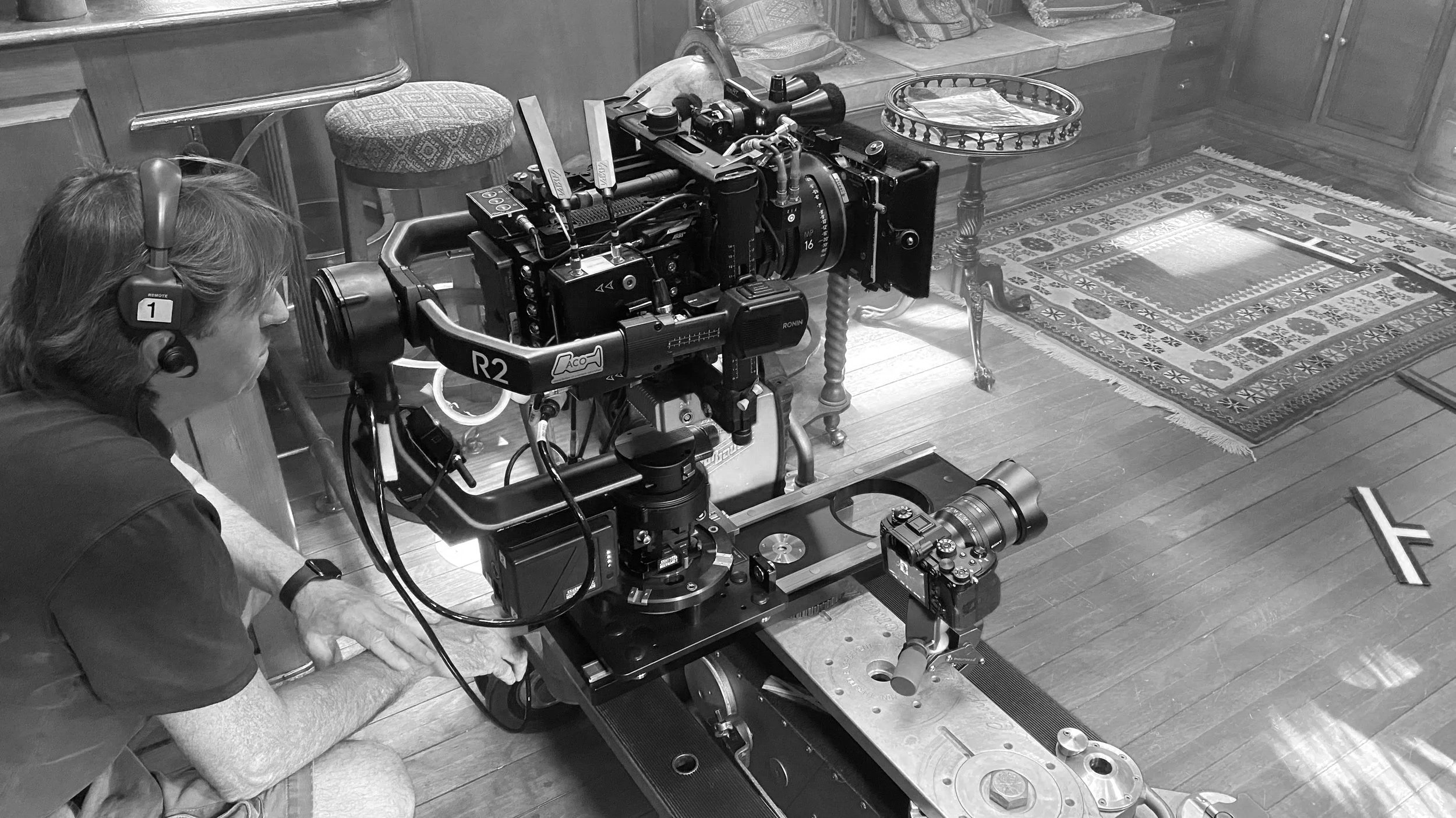 A person with headphones sitting on the floor beside a large camera rig on a dolly, capturing footage in a room with a patterned rug and wooden furniture.