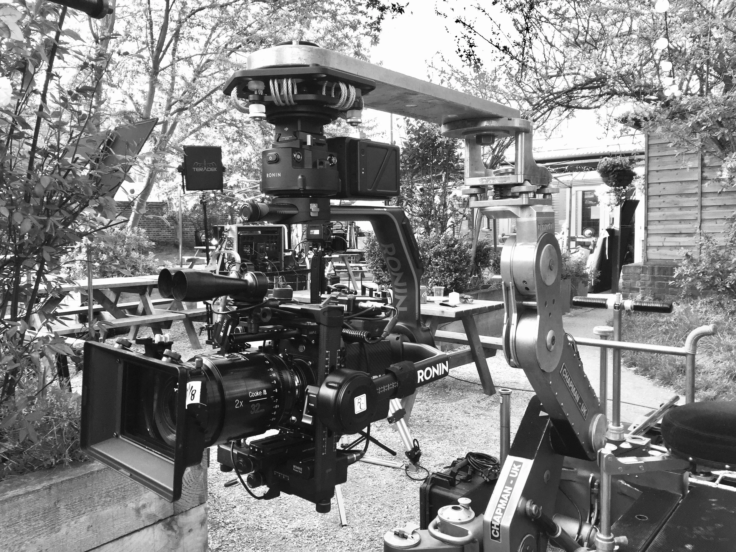 Black and white photograph of a camera mounted on a professional camera dolly, with trees, bushes, and a wooden structure in the background.