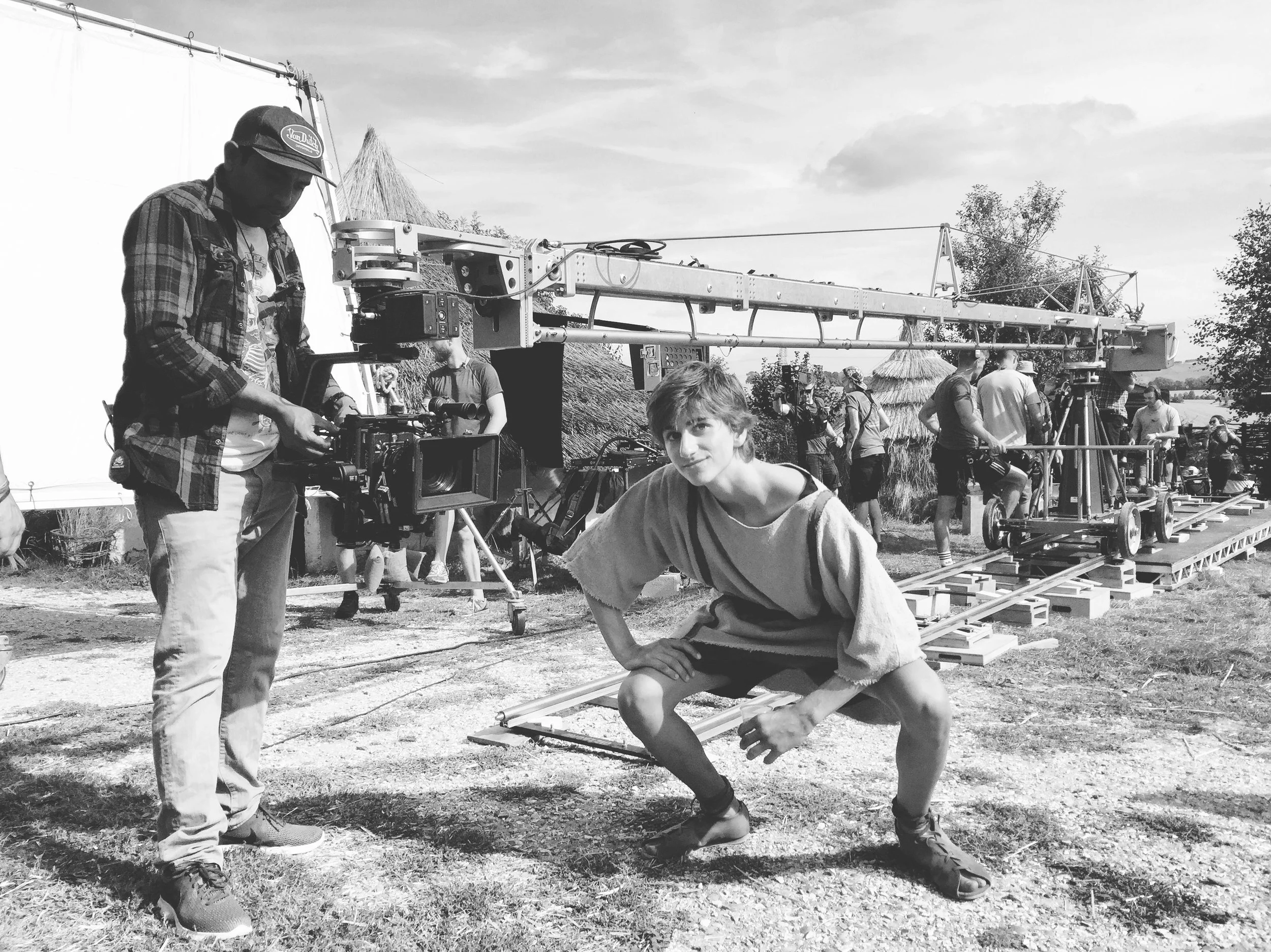 A film crew on outdoor set with camera assistant, young actor crouched in front, and crew members in the background handling equipment and lighting. The scene is in a rural area with trees and straw huts.