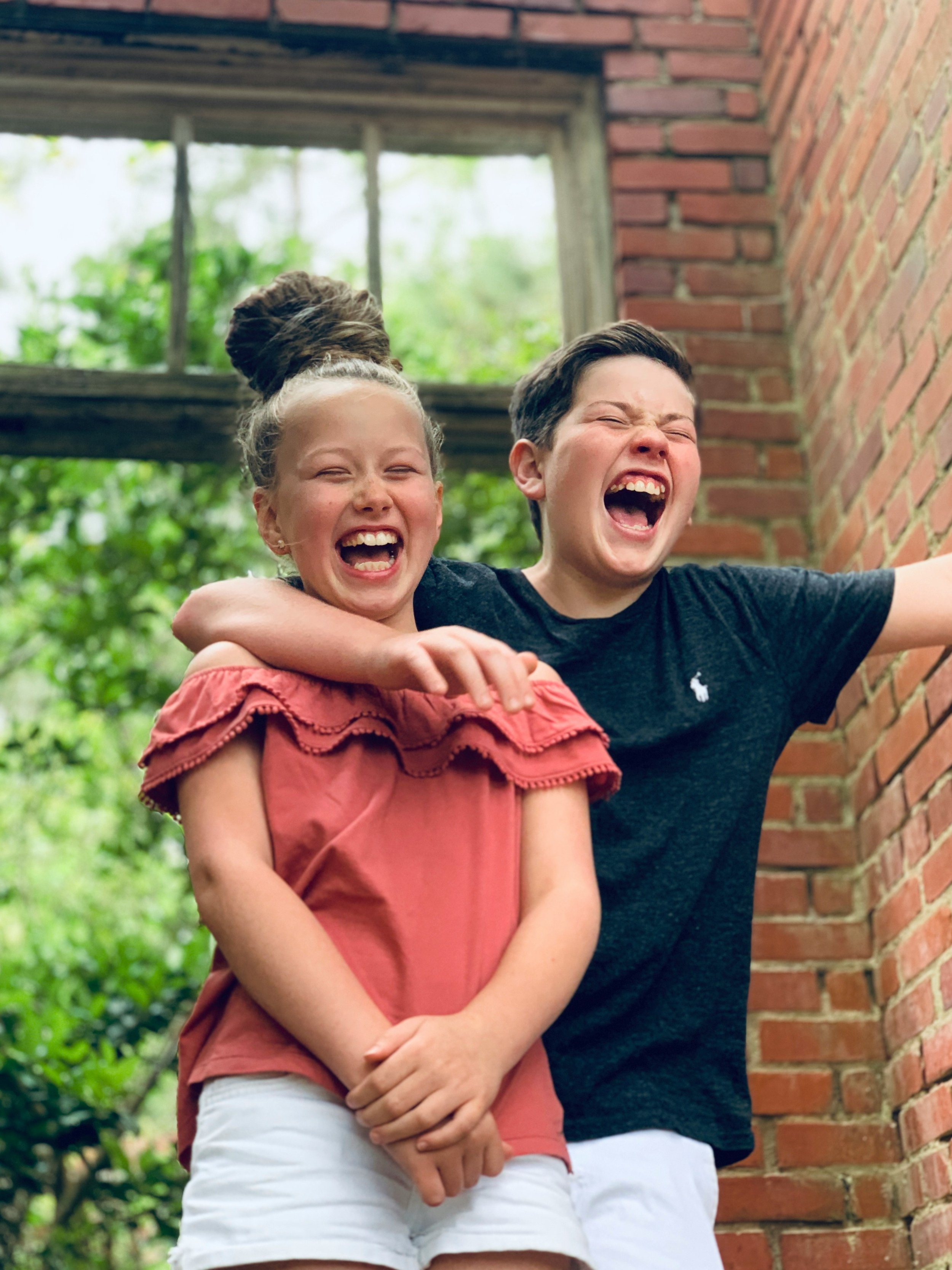 Two children, a girl and a boy, are laughing and hugging outside near a brick wall, with greenery and trees in the background.