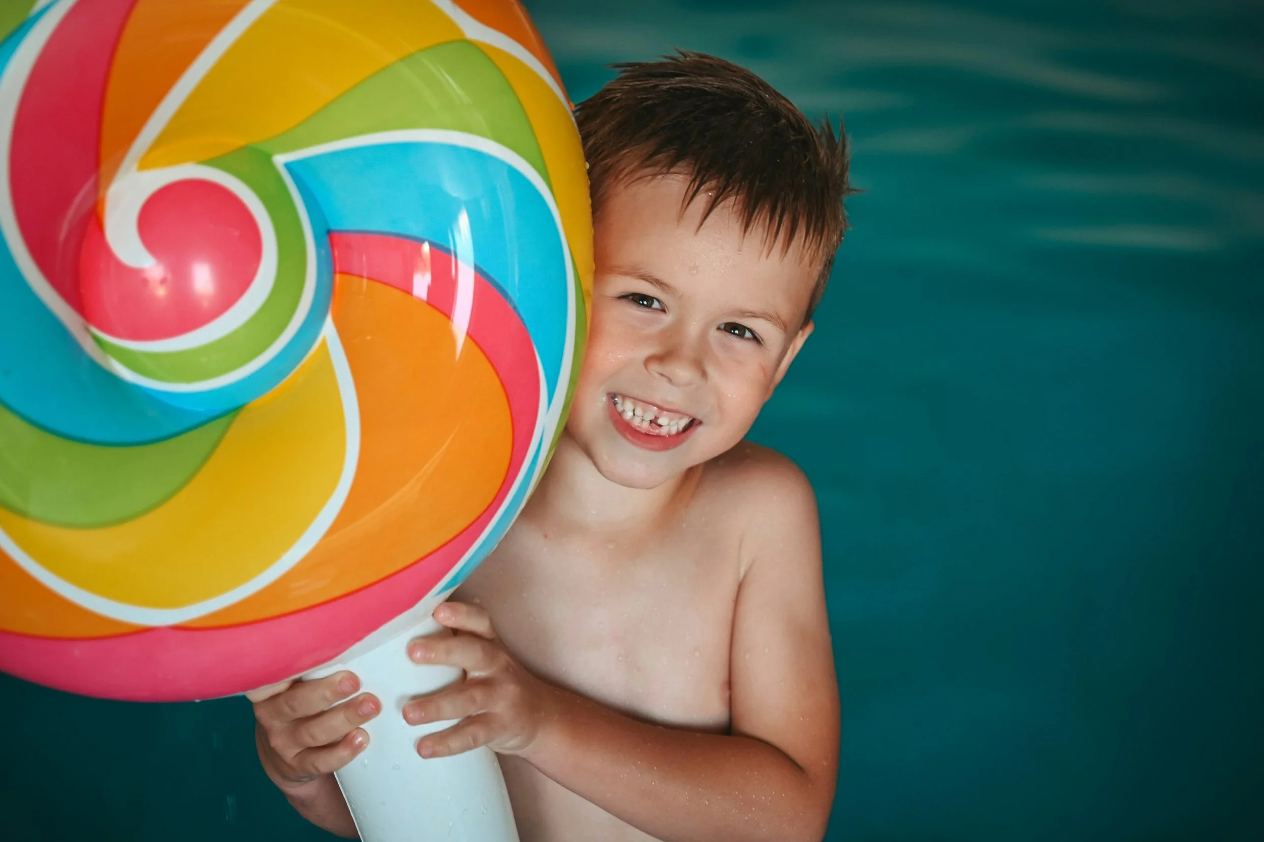 A young boy smiling in a swimming pool, holding a colorful spiral-patterned beach ball next to his face.