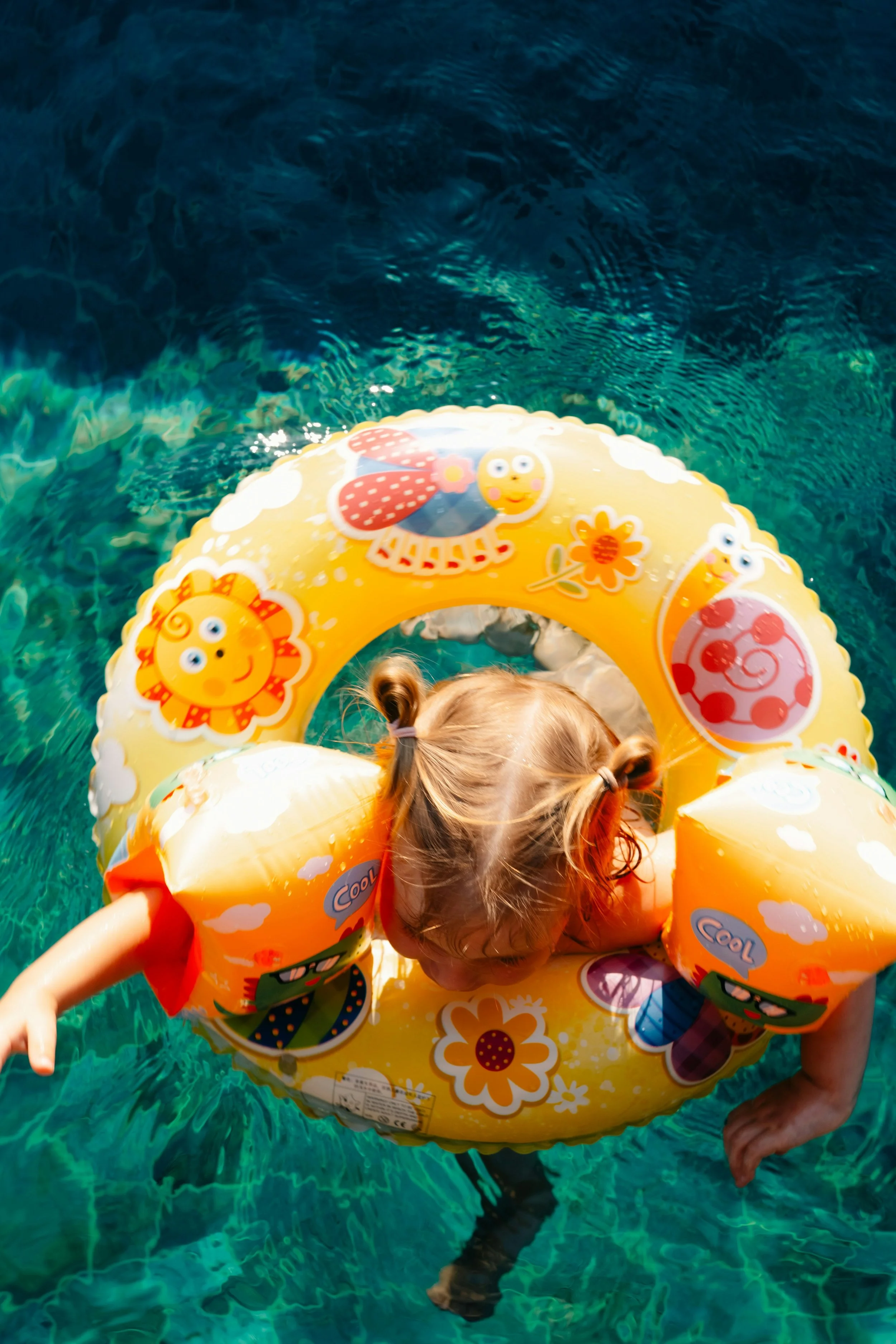 Child with two pigtails wearing a yellow flotation ring with cartoon animal designs, swimming in a pool with clear blue water.