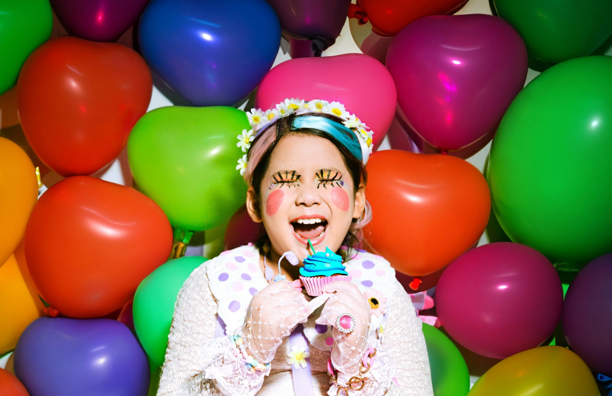A young girl with face paint resembling a clown, wearing a floral headband, holding a cupcake with blue frosting, surrounded by colorful balloons.