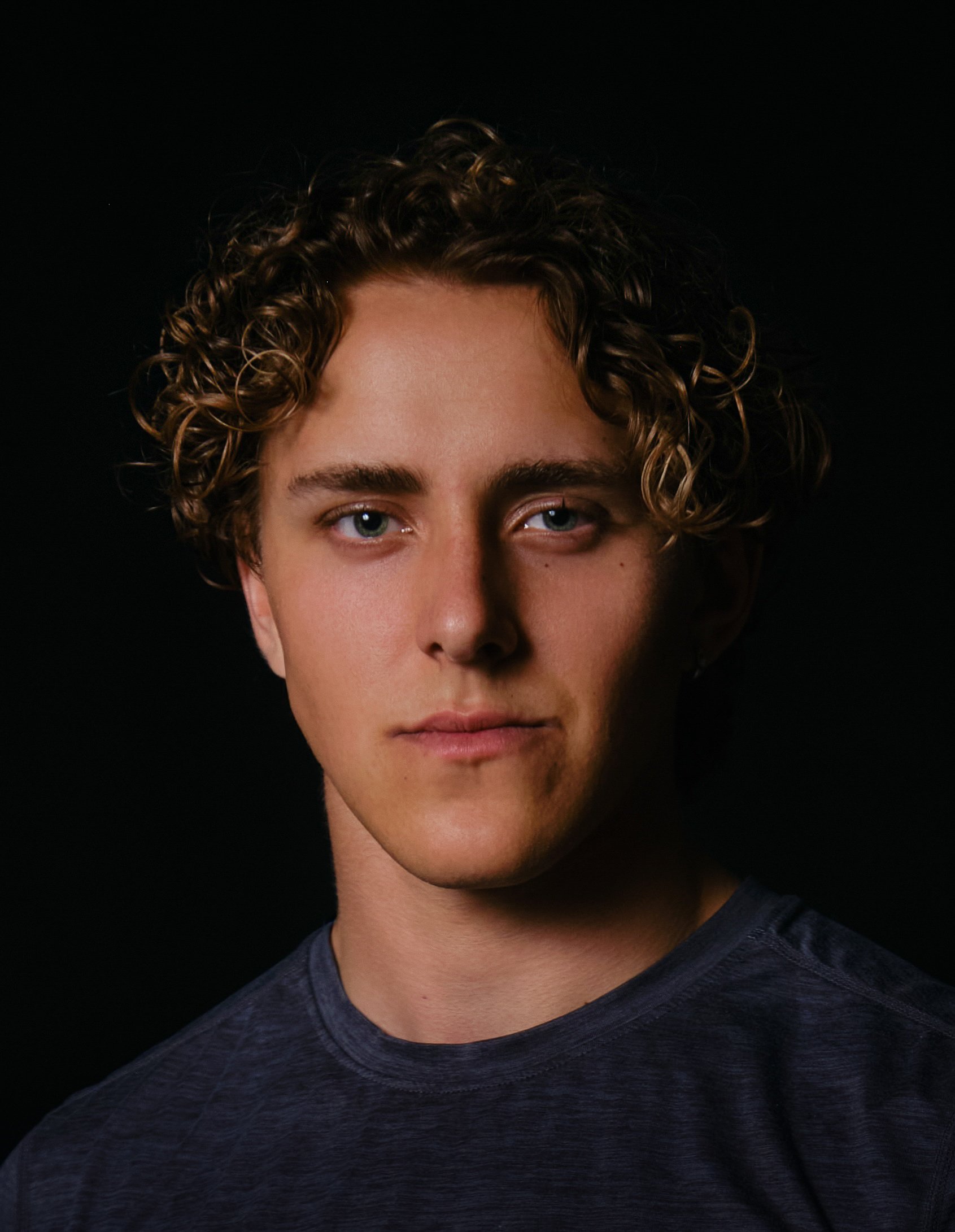 Close-up of a young man with curly brown hair and blue eyes, wearing a dark t-shirt, against a black background.